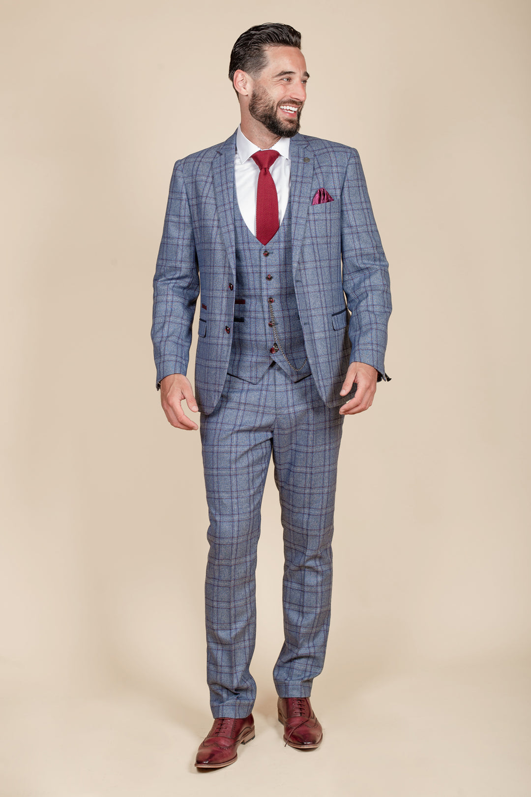 A man with short dark hair and a beard smiles in a marcdarcy ABBOTT blue tweed check three piece suit, styled with a white shirt, red tie, red pocket square, and brown dress shoes against a beige background.