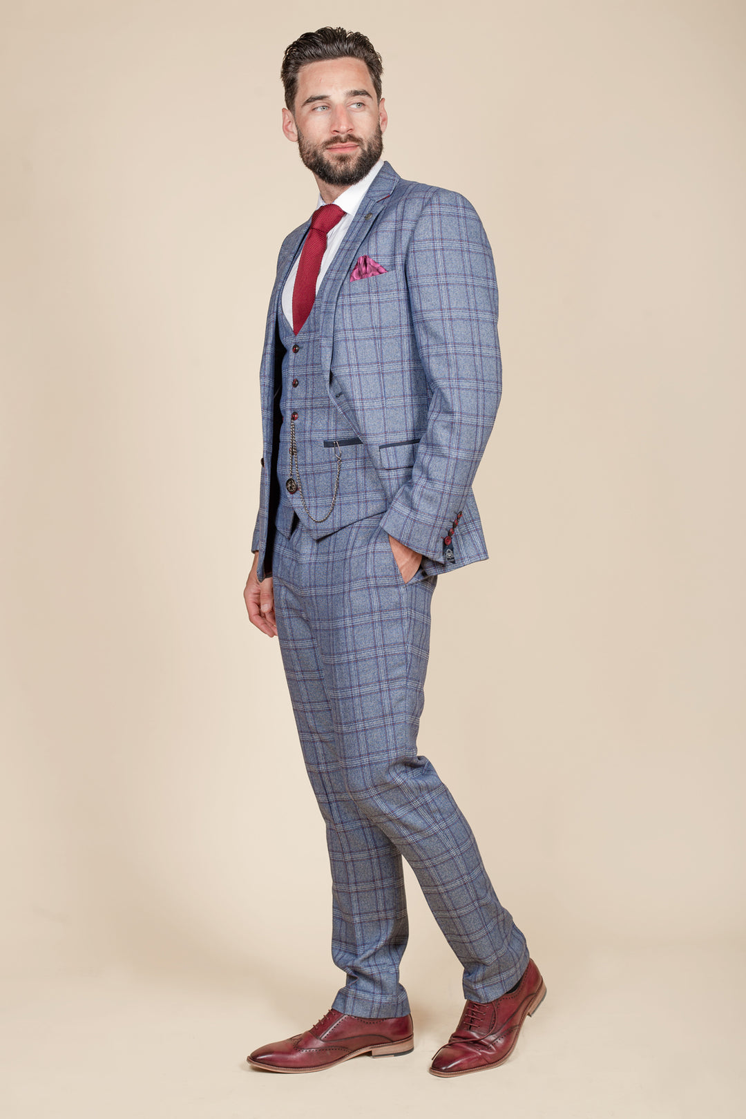 A bearded man stands against a beige background, wearing the marcdarcy ABBOTT Blue Tweed Check Three Piece Suit with a red tie, matching pocket square, and red shoes, looking to the side with one hand in his pocket.