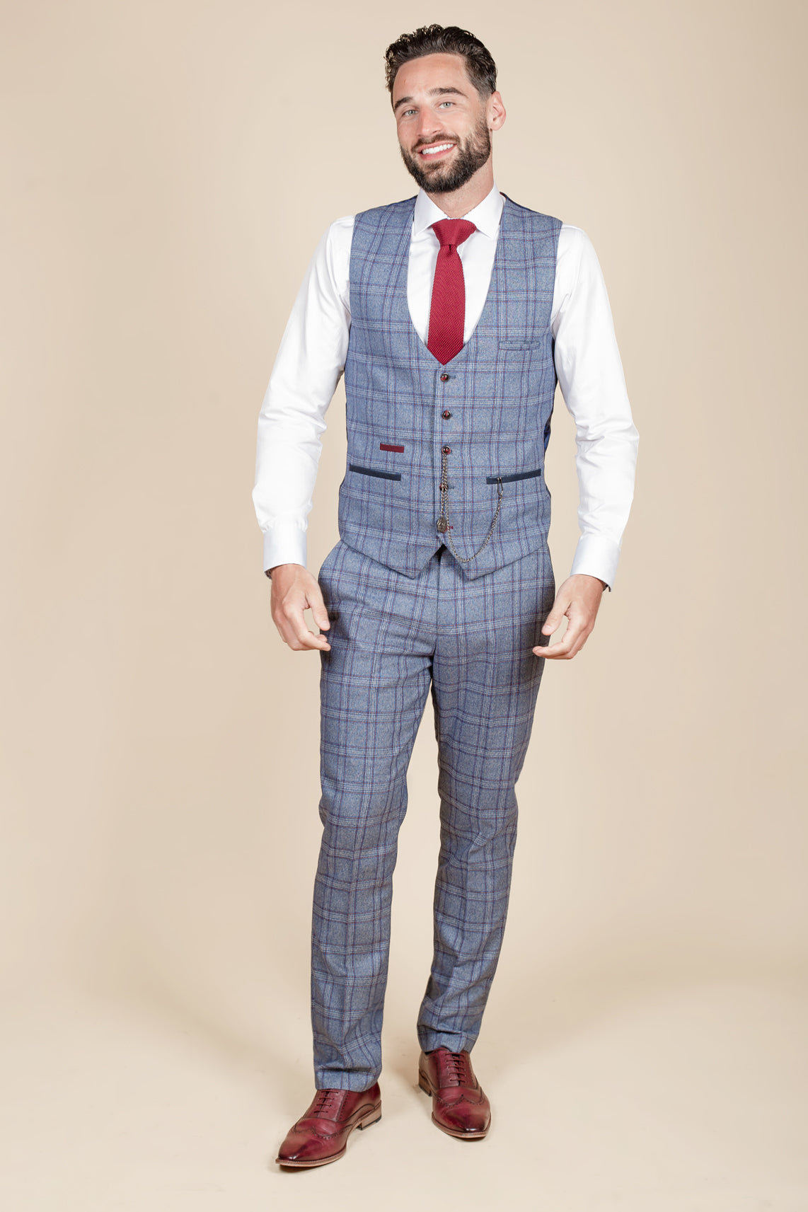 A man with short brown hair and a beard smiles while wearing the marcdarcy ABBOTT Blue Tweed Check Waistcoat, a white shirt, red tie, and brown dress shoes. He stands against a plain beige background.