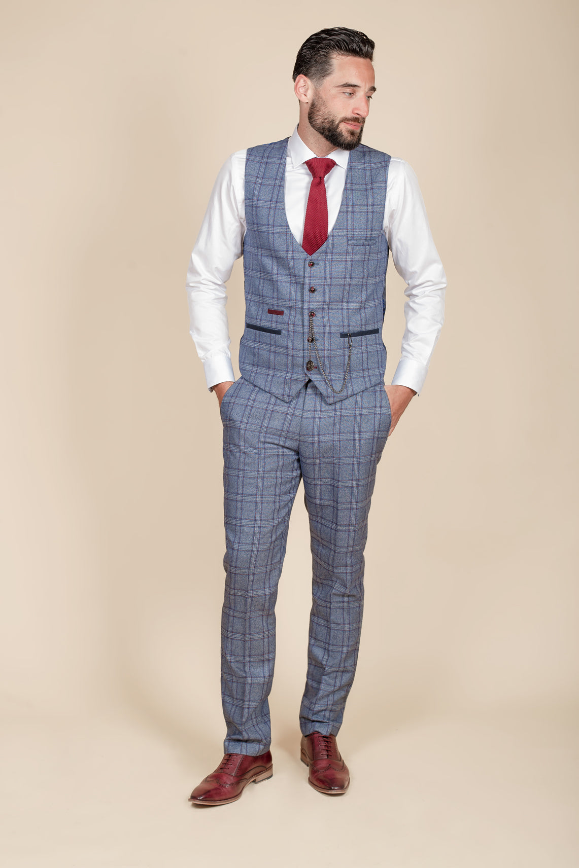 A man with dark hair and a beard models the marcdarcy ABBOTT Blue Tweed Check Waistcoat over a white shirt and red tie, embodying contemporary heritage style as he stands hands-in-pockets against a cream background.