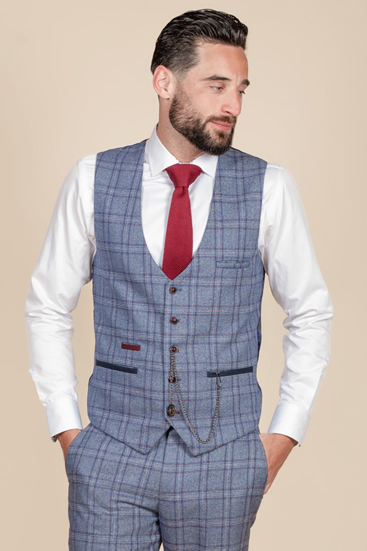 A dark-haired, bearded man wears the marcdarcy ABBOTT Blue Tweed Check Waistcoat with matching trousers, a white shirt, and red tie. Hands in pockets, he gazes left against a beige backdrop, channeling contemporary heritage style.