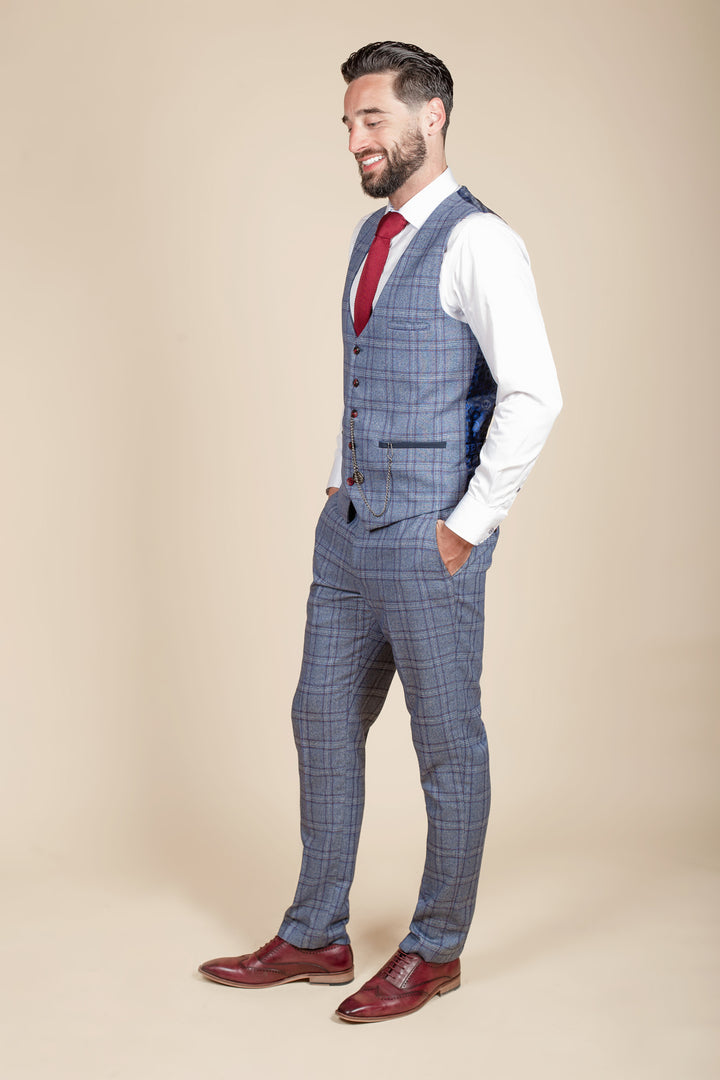A man with dark hair and a beard smiles in profile, wearing the marcdarcy ABBOTT Blue Tweed Check Waistcoat over a blue checked suit, with a red tie, white shirt, and red shoes, hands in pockets against a beige background.