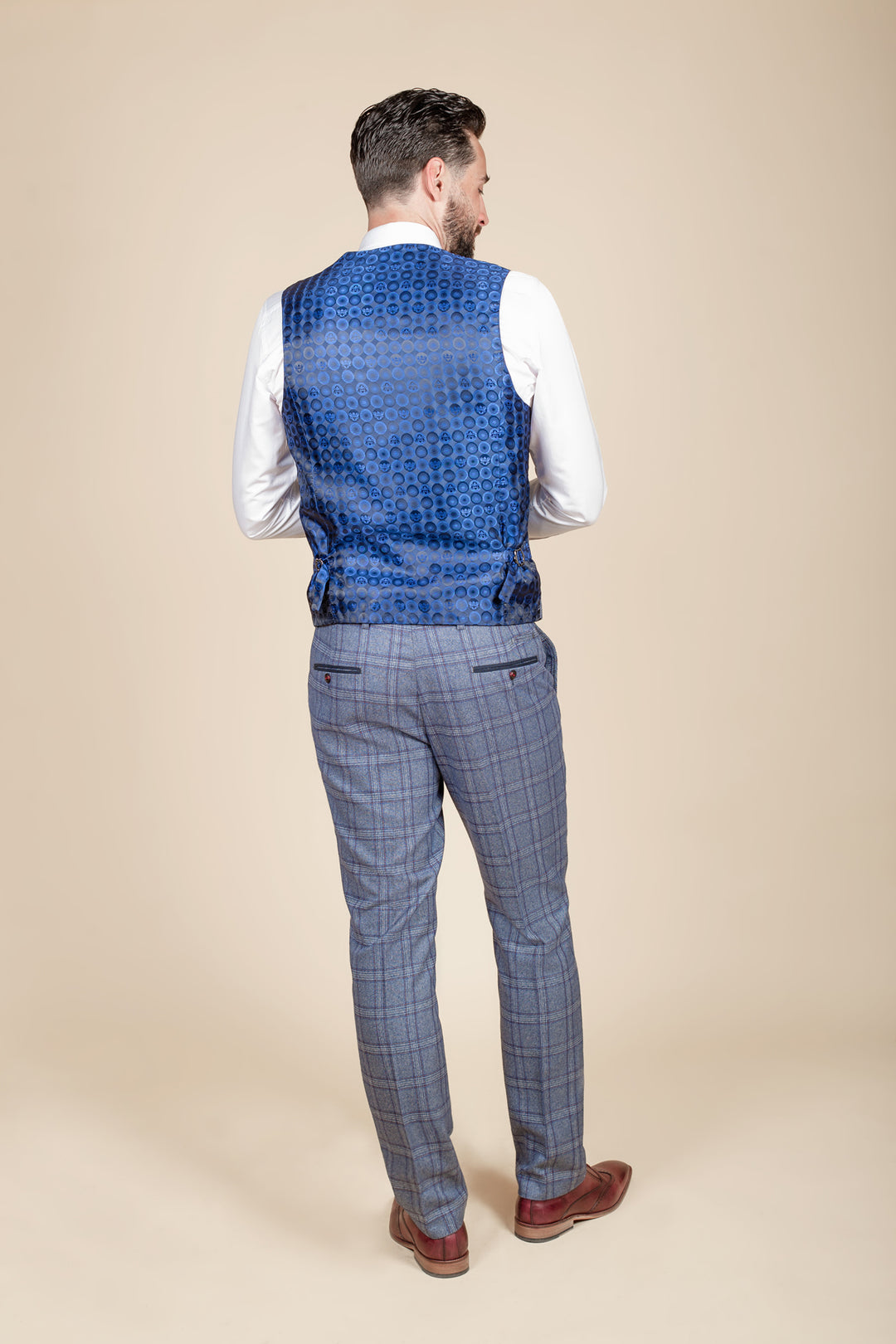 A man faces away, wearing the marcdarcy ABBOTT Blue Tweed Check Waistcoat over a white shirt, gray plaid trousers, and brown shoes, set against a plain beige background.