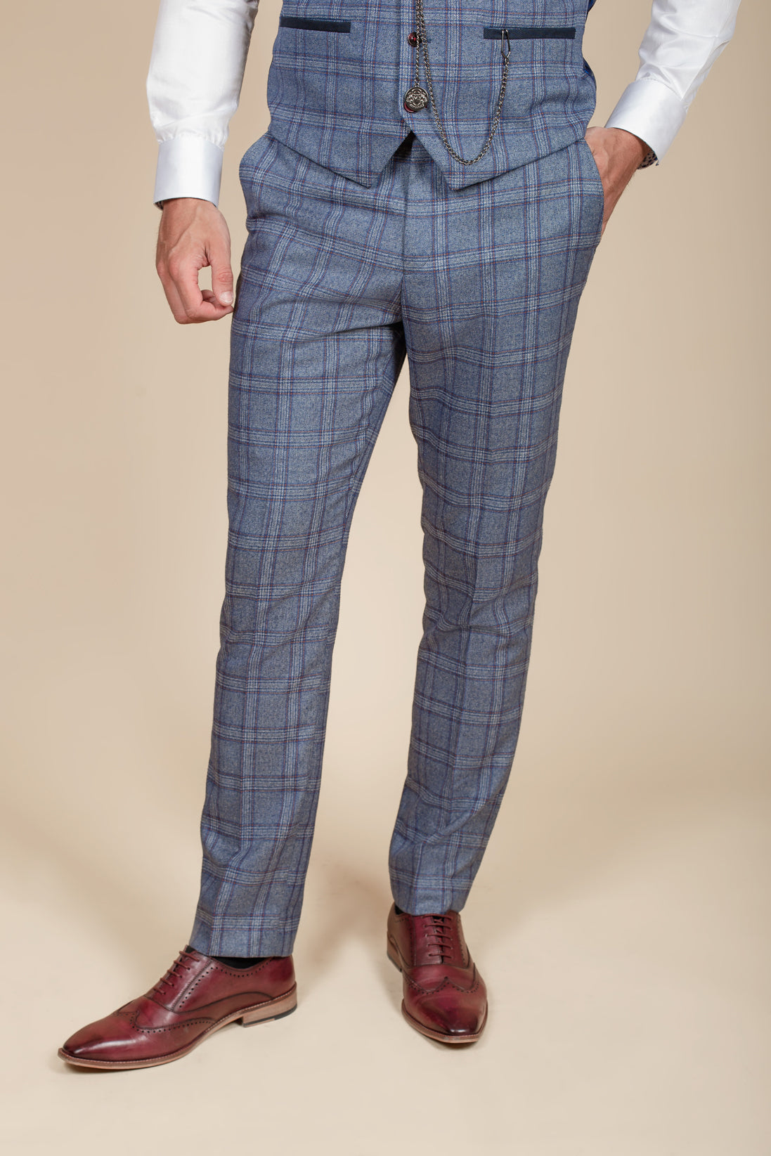 A person wearing the marcdarcy ABBOTT Blue Tweed Check Three Piece Suit stands with one hand in their pocket against a beige background. Their face is not visible, and they pair the suit with a white shirt and red leather dress shoes.