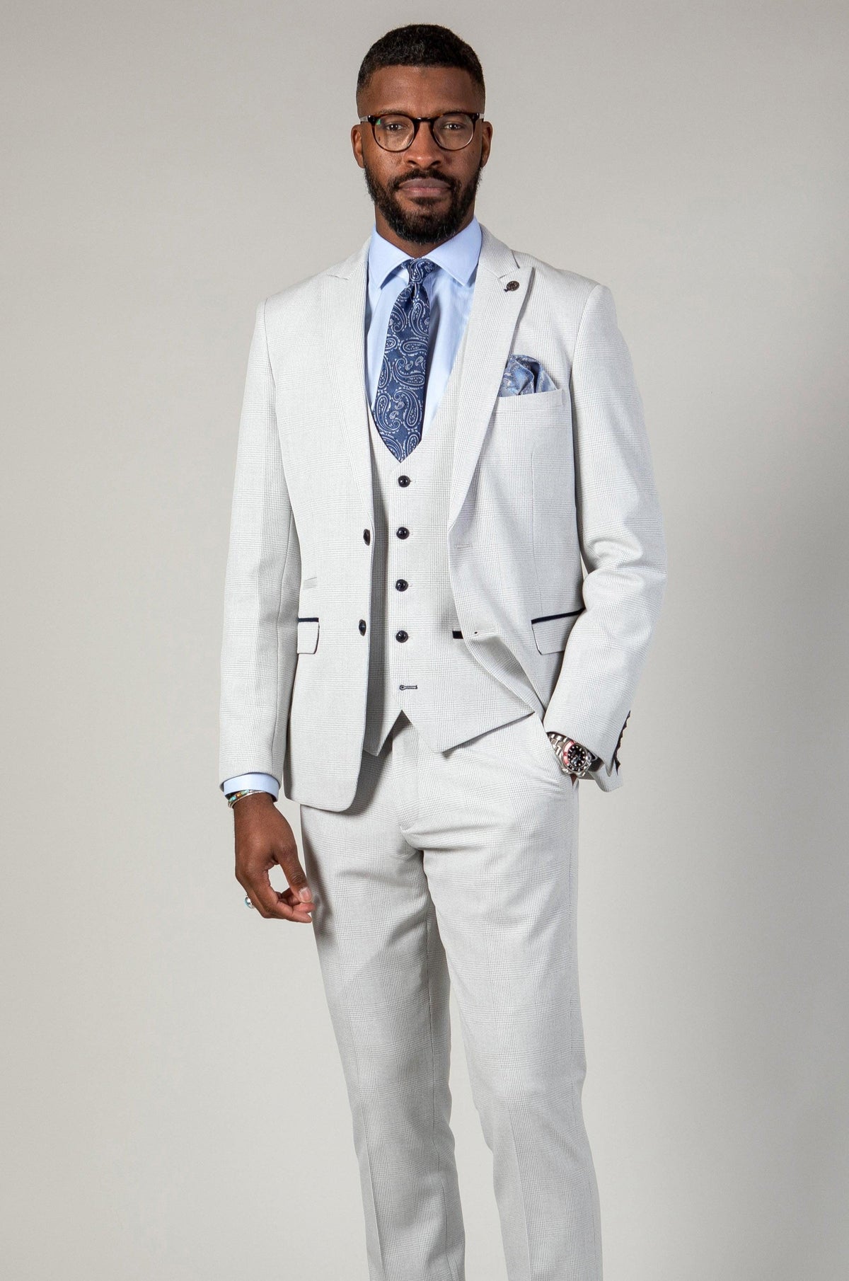 A man wearing the Marc Darcy WBA Collection BROMLEY Stone Suit with a Kelvin Navy Waistcoat, paired with a blue patterned tie and pocket square, stands against a light gray background. He has short hair, glasses, and a trimmed beard.