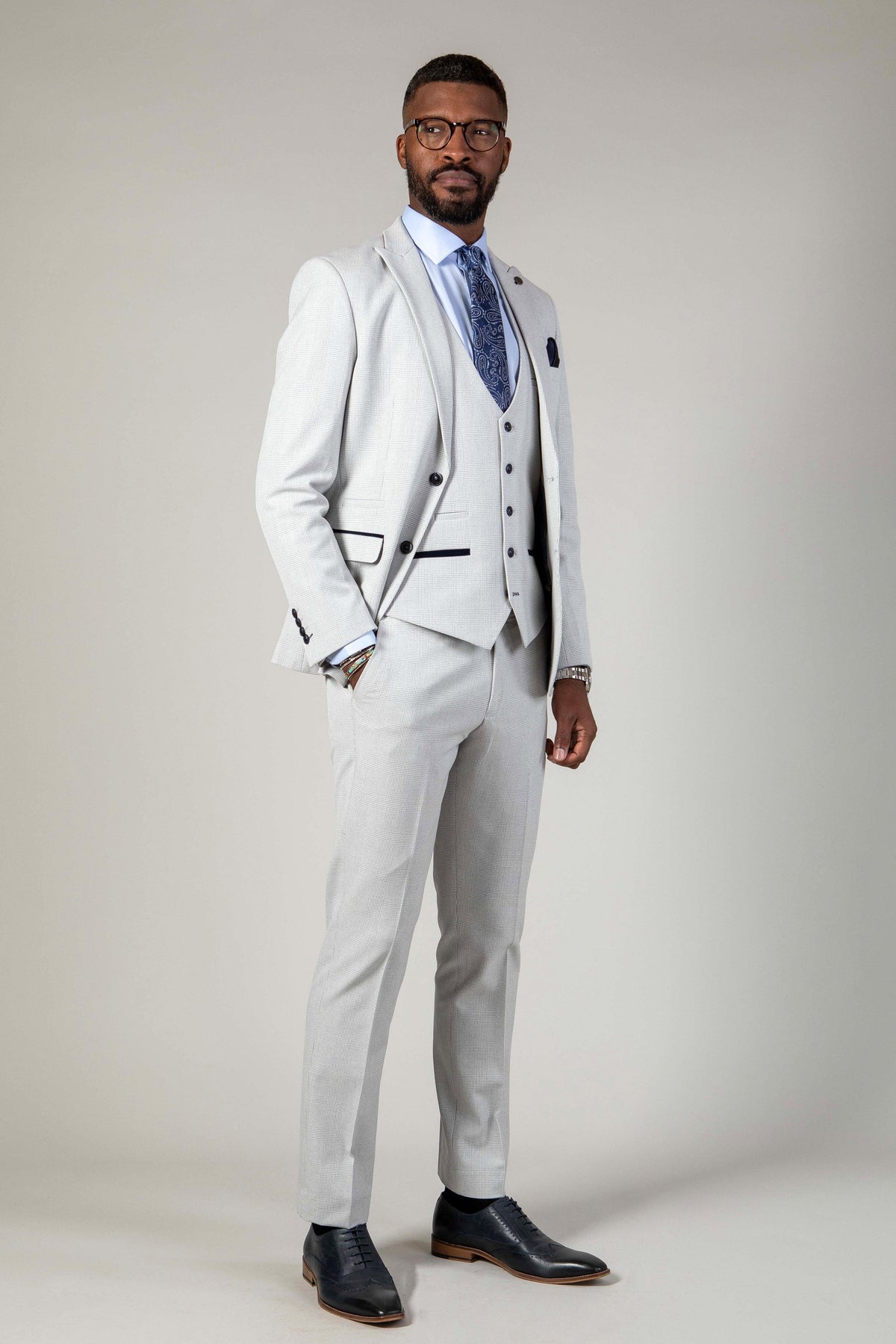 A man stands confidently in Marc Darcy's The WBA Collection - BROMLEY Stone Suit with Kelvin Navy Waistcoat, styled with a white shirt, patterned blue tie, and dark dress shoes against a plain background.
