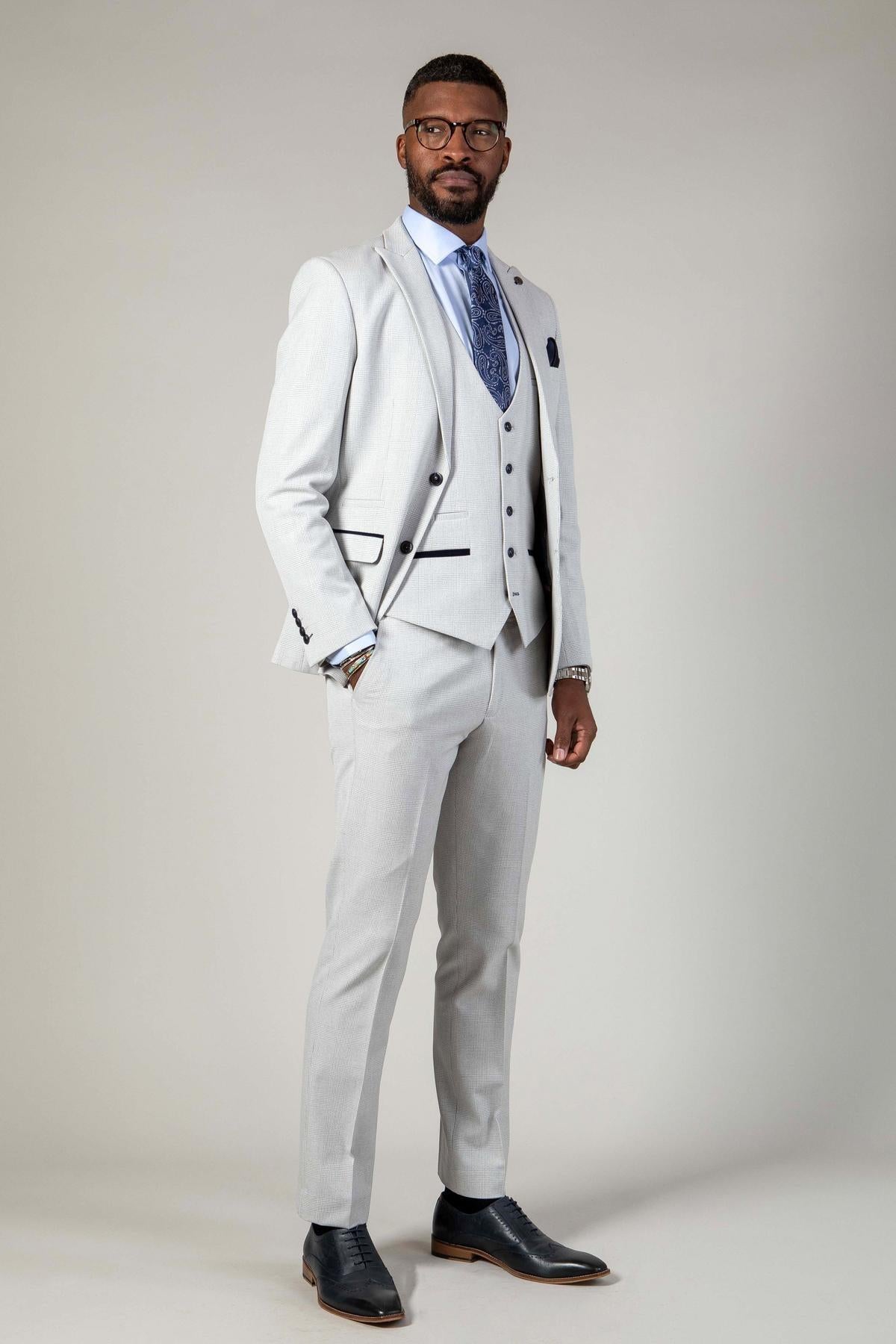 A man stands confidently in the Marc Darcy WBA Collection - BROMLEY Stone Suit, styled with a blue patterned tie and black shoes. He has short hair, a beard, and glasses, set against a plain background.