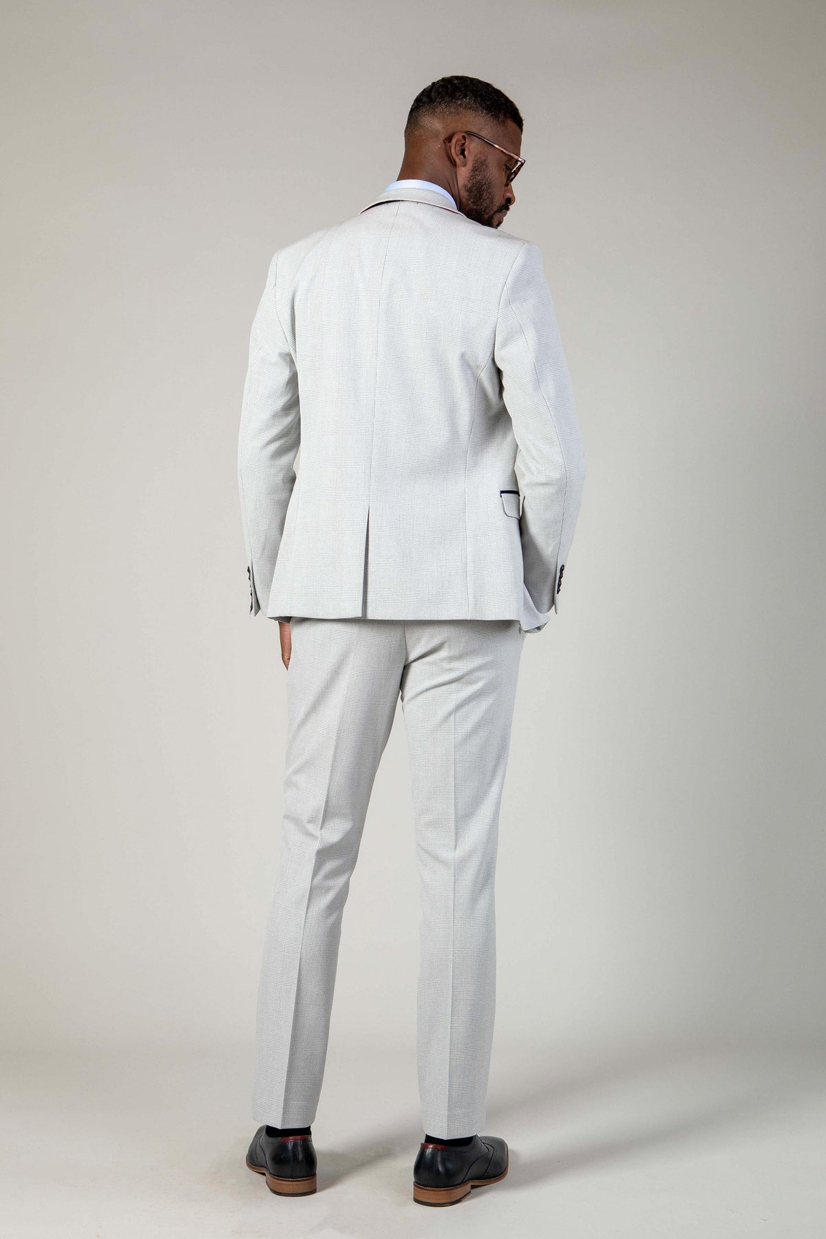 A man in the Marc Darcy WBA Collection BROMLEY Stone Suit with Kelvin Navy Waistcoat and black shoes stands facing away from the camera against a plain, light background.