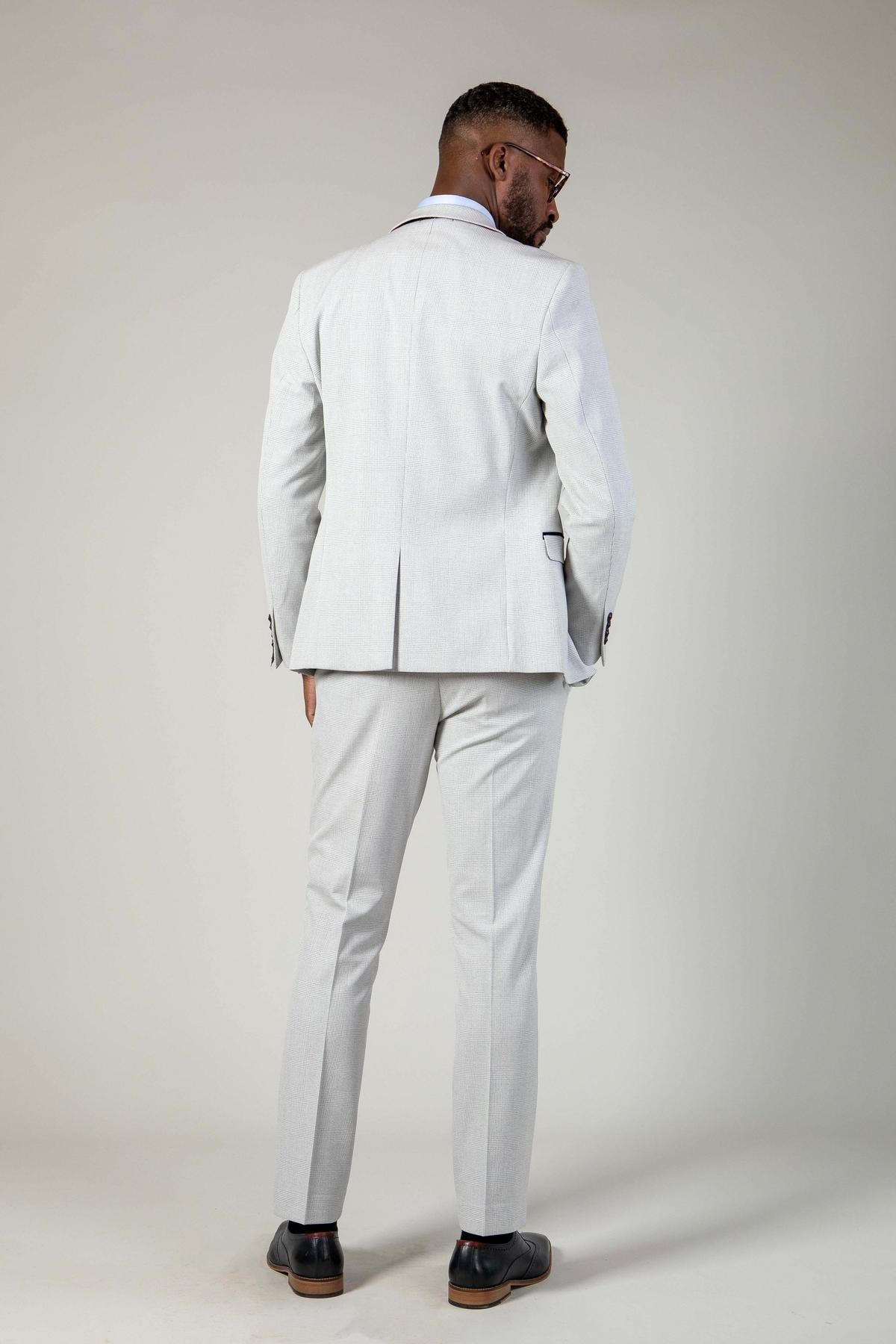 A man in the Marc Darcy BROMLEY Stone Suit from The WBA Collection stands facing away, hands in pockets, wearing dark shoes and glasses against a plain light background.