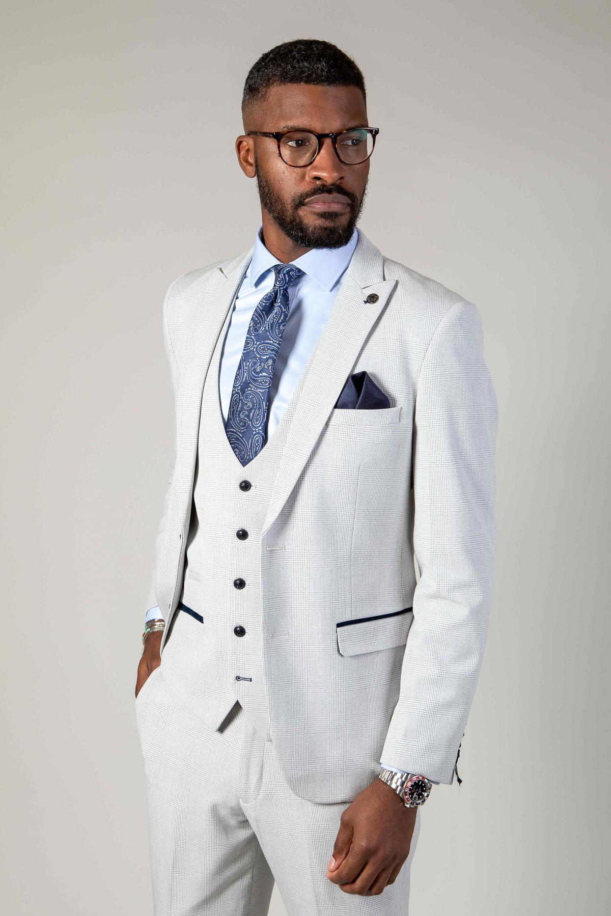 A man wearing the Marc Darcy WBA Collection - BROMLEY Stone Suit with black buttons, a white shirt, blue patterned tie, and dark-rimmed glasses stands hands-in-pocket, looking to the side against a plain background.
