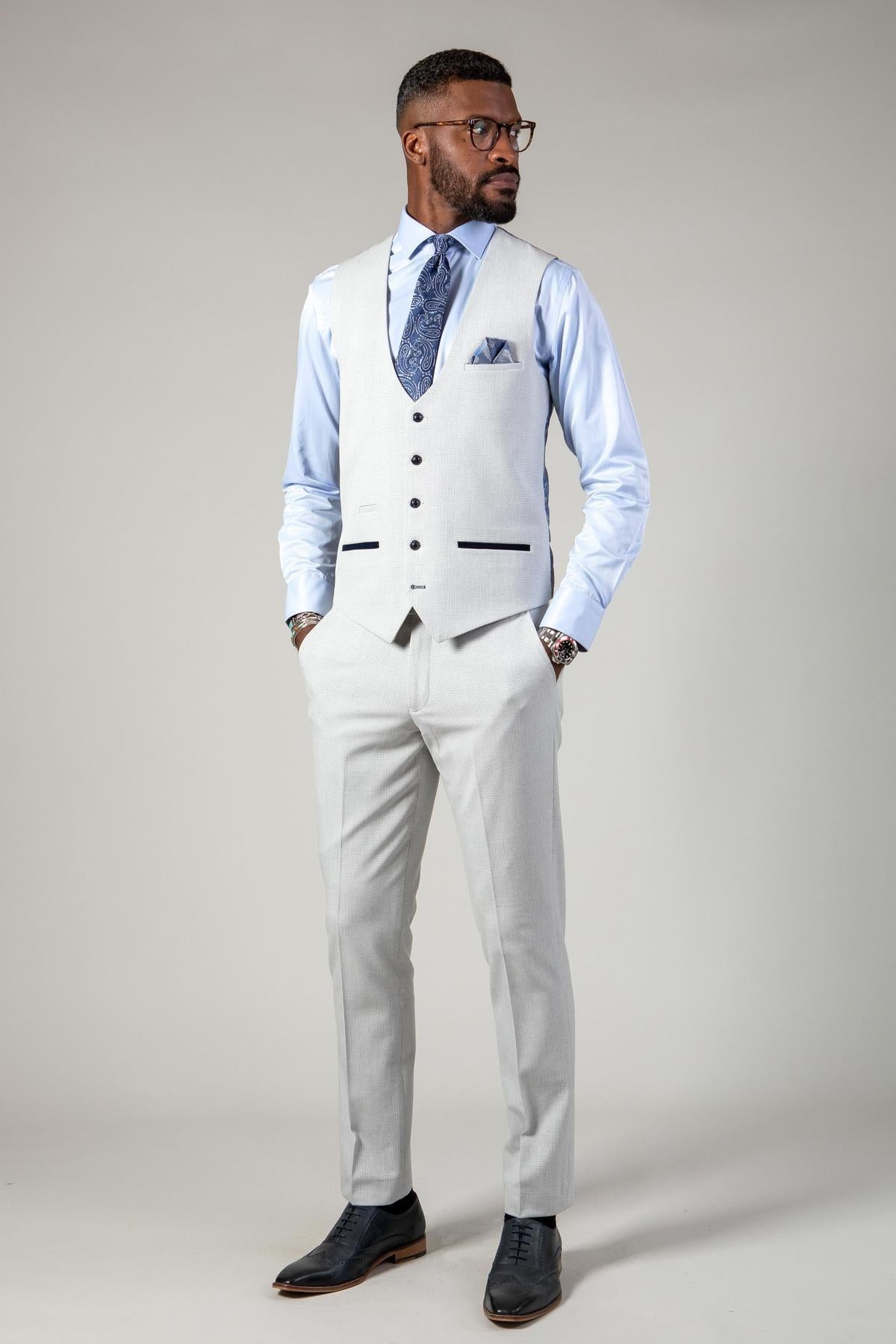 A man wearing glasses stands confidently in the Marc Darcy WBA Collection - BROMLEY Stone Suit, featuring a Prince of Wales check three-piece set with vest, light blue shirt, patterned tie, and black shoes against a gray background.