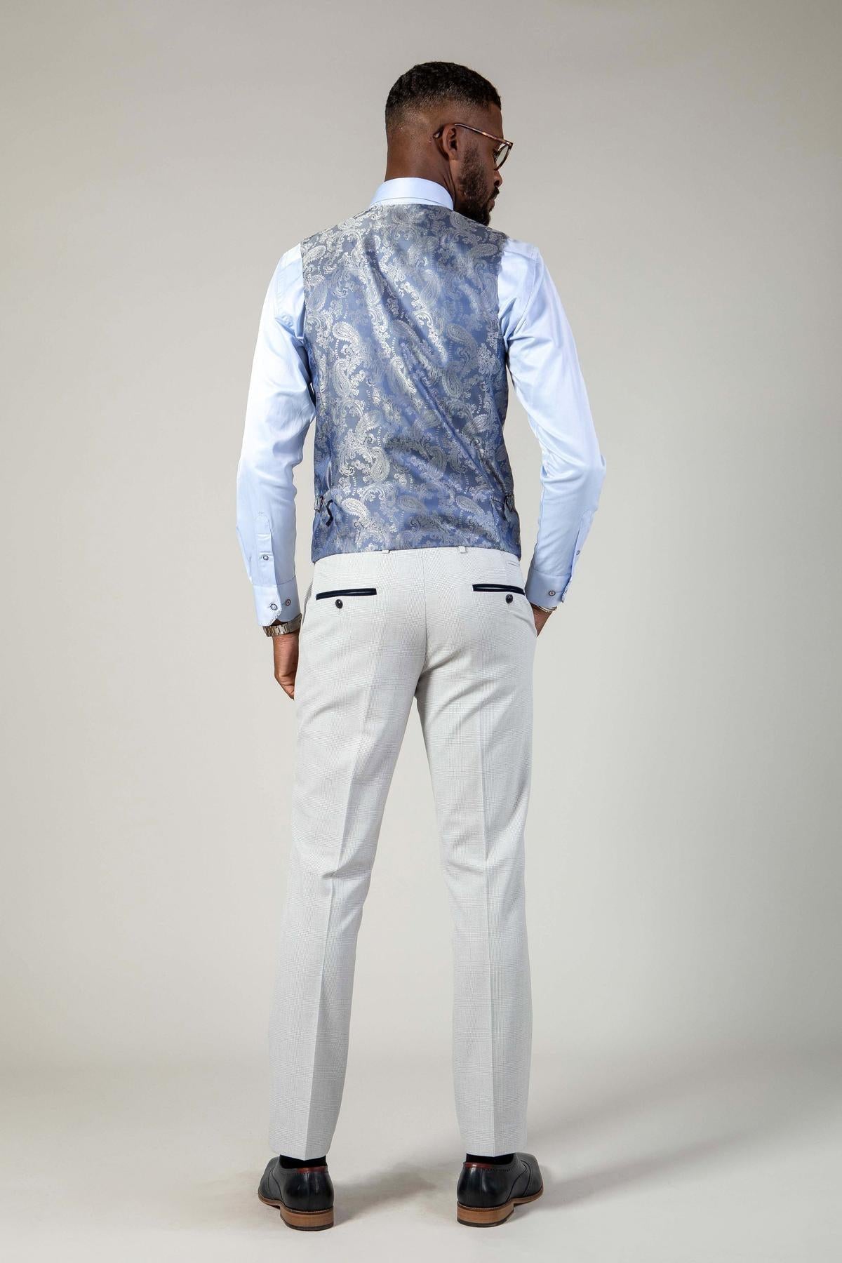A man stands facing away, wearing the Marc Darcy BROMLEY Stone Suit from The WBA Collection, featuring a light blue shirt and waistcoat, light gray dress pants, and black dress shoes against a plain light background.