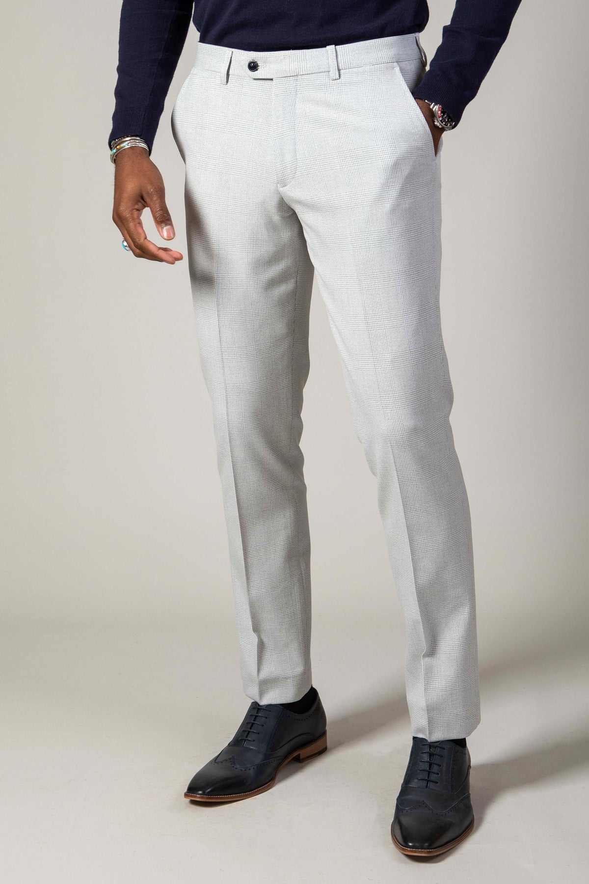 A man wears Marc Darcy’s The WBA Collection – BROMLEY Stone Suit trousers with a navy Kelvin waistcoat, styled with a navy sweater and dark oxford shoes. The flat-front trousers offer belt loops and single button closure for a refined look.