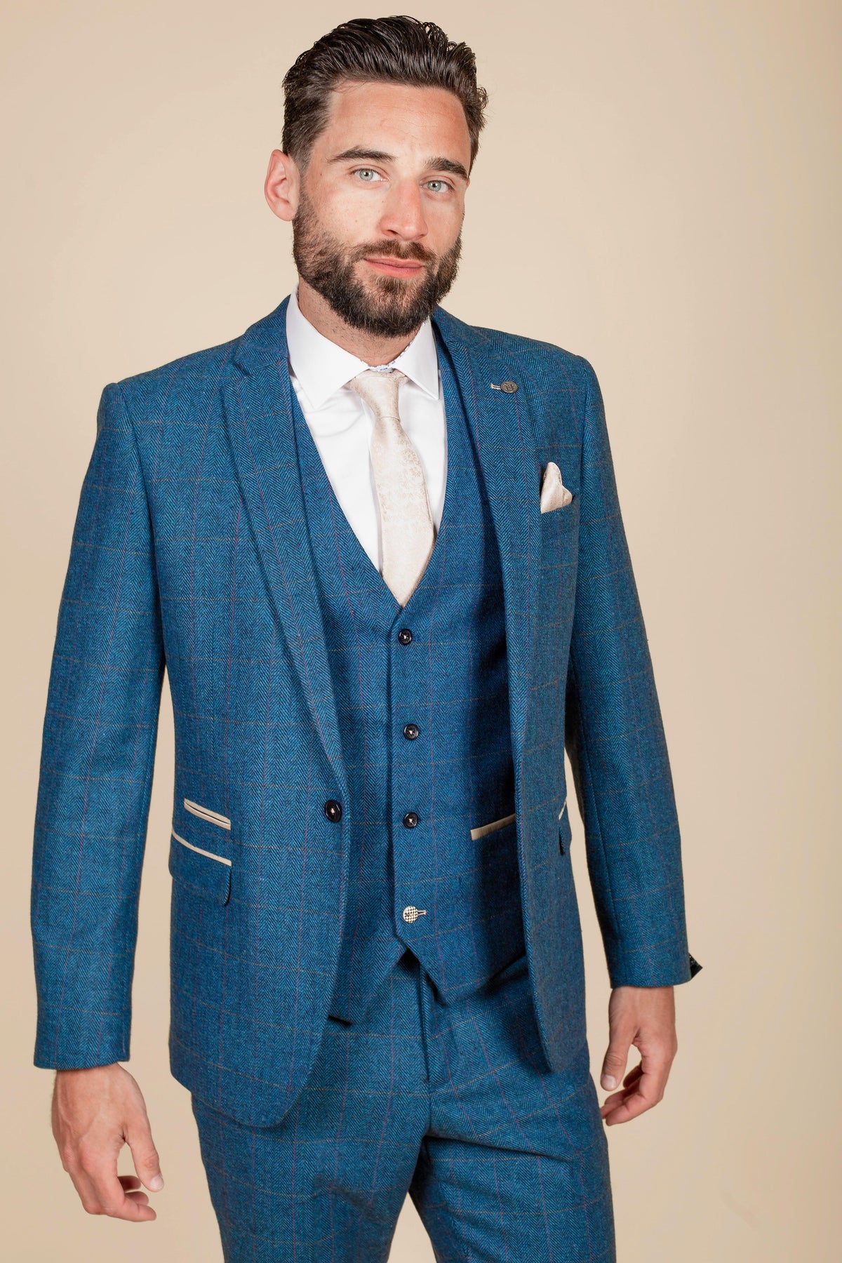 A man with dark hair and a beard is wearing the marcdarcy Everton Collection DION Blue Tweed Suit with the Kelvin waistcoat, paired with a white shirt and beige tie, standing against a plain beige background.