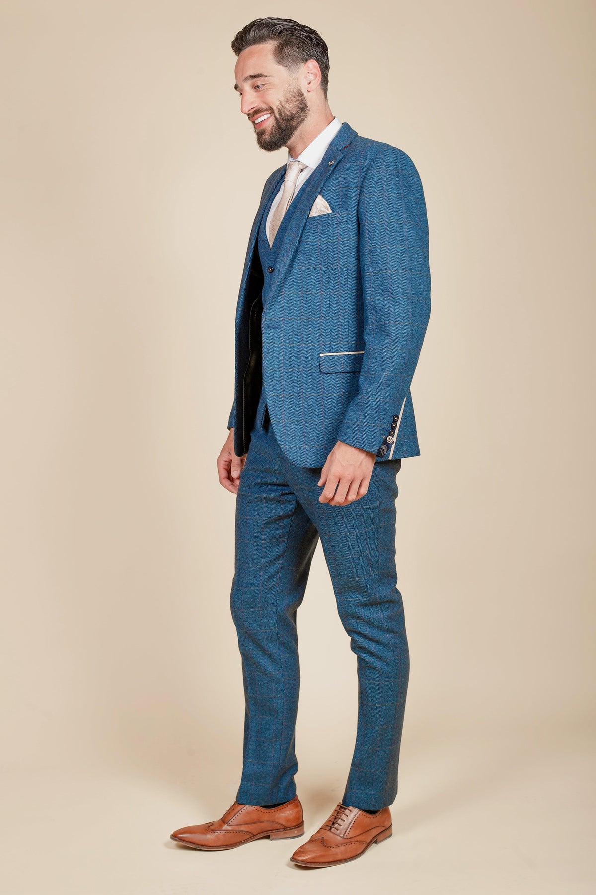 Jarrad Branthwaite stands smiling in the marcdarcy Everton Collection, wearing a DION Blue Tweed Check Suit, white shirt, light tie, brown dress shoes, and beige pocket square against a plain beige background.
