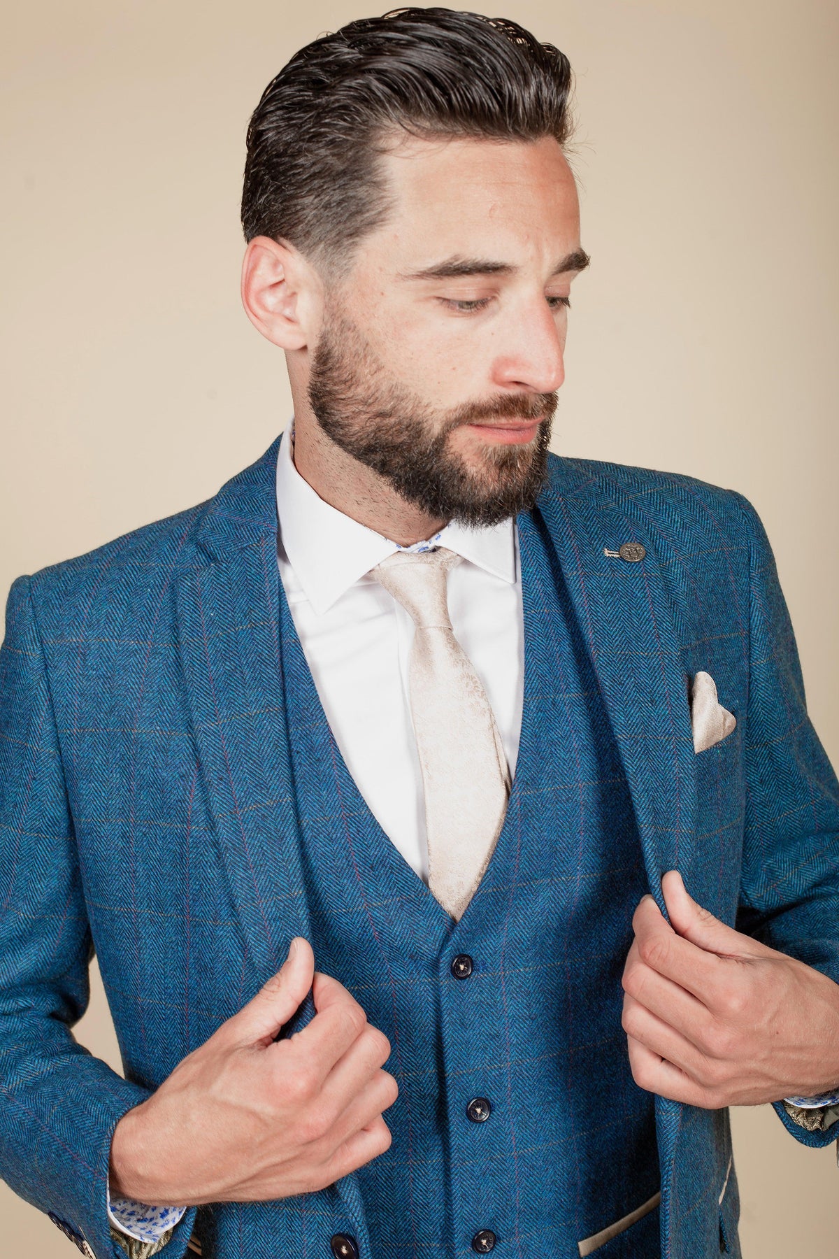 A man with dark hair and a beard wears marcdarcy’s The WHU Collection - DION Blue Tweed Suit with TED Waistcoat as worn by Crysencio Summerville, styled with a white shirt, beige tie, and matching pocket square. He looks down while holding his jacket.