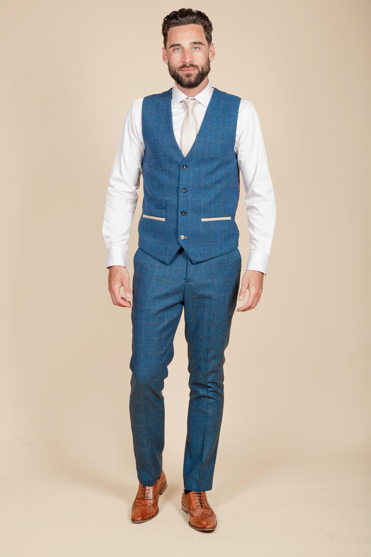 A man wears the marcdarcy Everton Collection Séamus Coleman DION Blue Tweed Check Suit, featuring a blue plaid vest, white shirt, and tan tie, standing against a plain beige background.