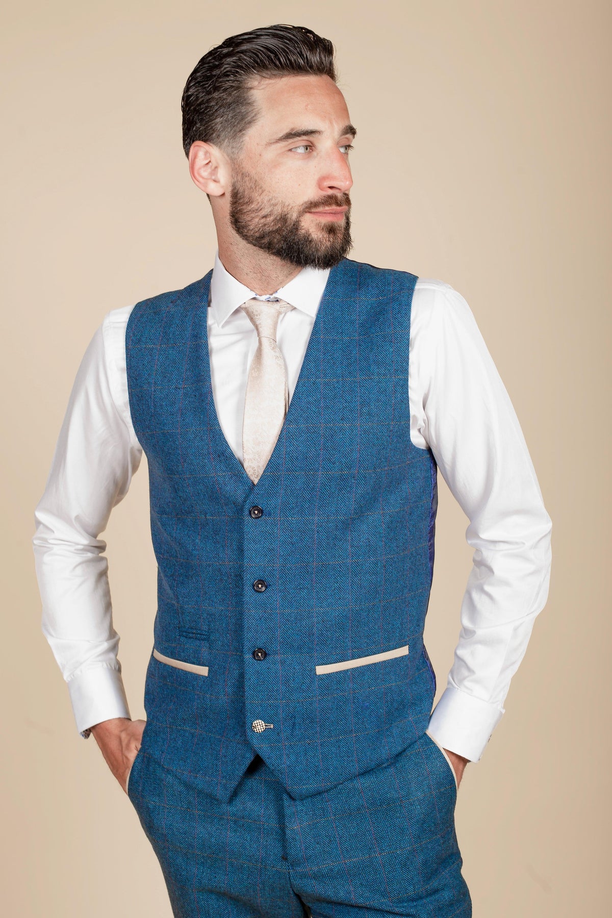 A man with dark hair and a beard models the marcdarcy WHU Collection - DION Blue Tweed Check Suit as worn by Crysencio Summerville, featuring a vest and trousers over a white shirt and tie, hands in pockets against a beige background.