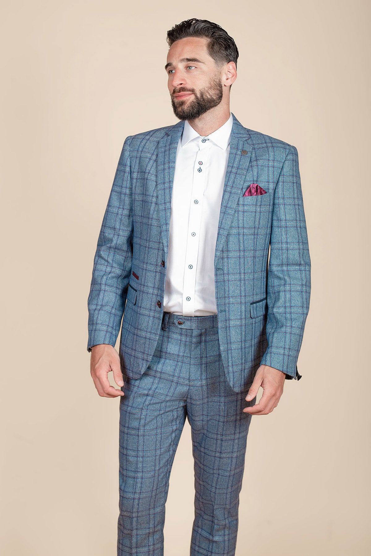 A dark-haired, bearded man models the marcdarcy ABBOTT Blue Tweed Check Blazer with a white shirt and magenta pocket square, set against a plain beige background for a modern heritage style look.