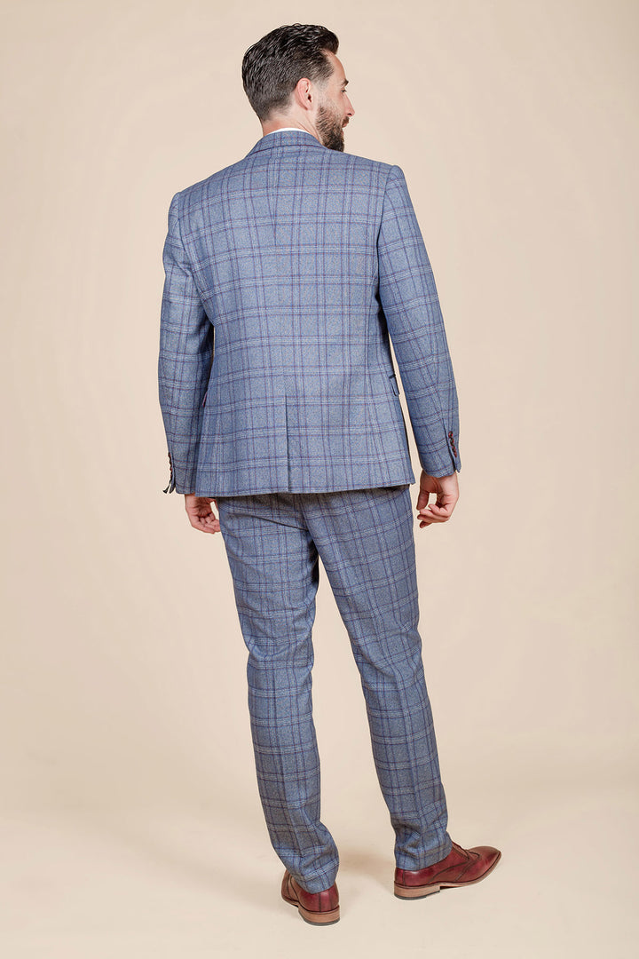 A man in a marcdarcy ABBOTT Blue Tweed Check Three Piece Suit stands with his back to the camera on a beige background, wearing brown dress shoes. He has short dark hair and a beard.