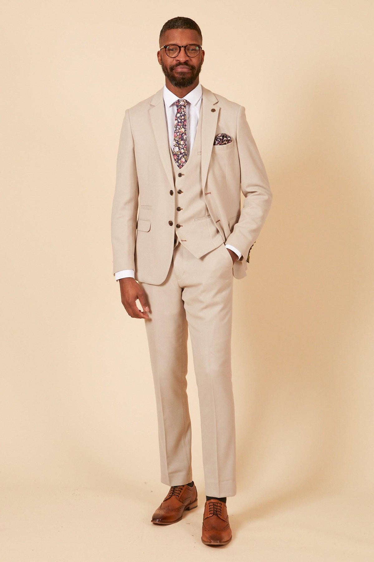 A man wears the Marc Darcy Leicester City F.C. Collection HM5 Stone Suit with Kelvin Black Waistcoat as worn by Mads Hermansen, styled with a floral tie, pocket square, glasses, and brown dress shoes.
