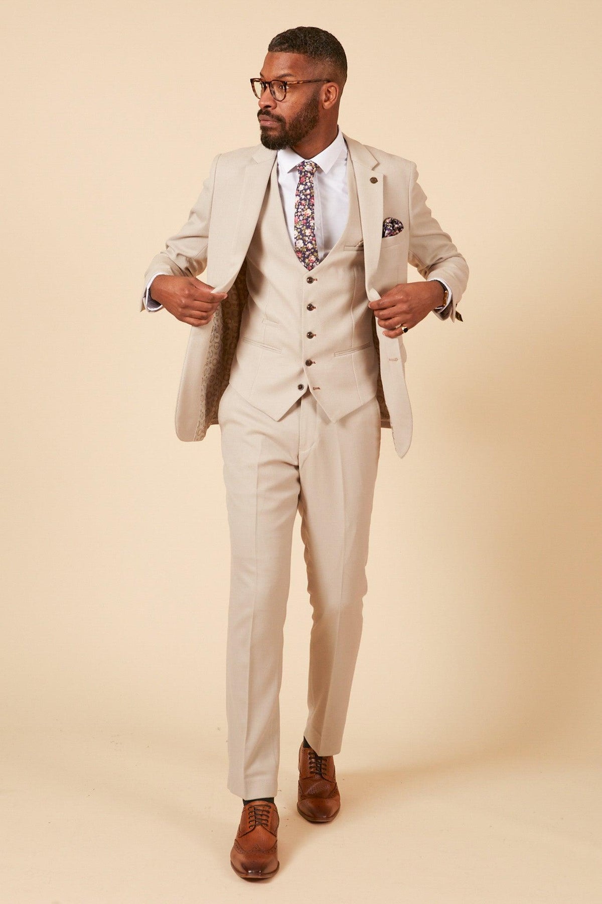 A man, adjusting his jacket and looking to the side, wears the Marc Darcy Leicester City F.C. Collection HM5 Stone Suit with Kelvin Black Waistcoat as worn by Mads Hermansen against a beige background.