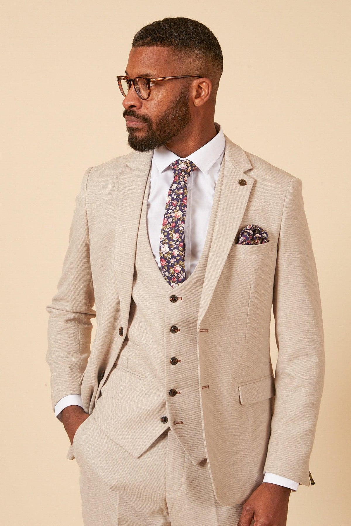 A man stands confidently in the Marc Darcy Leicester City F.C. Collection HM5 Stone Suit with Kelvin Black Waistcoat, inspired by goalkeeper Mads Hermansen, styled with glasses, a floral tie, pocket square, and a trimmed beard.