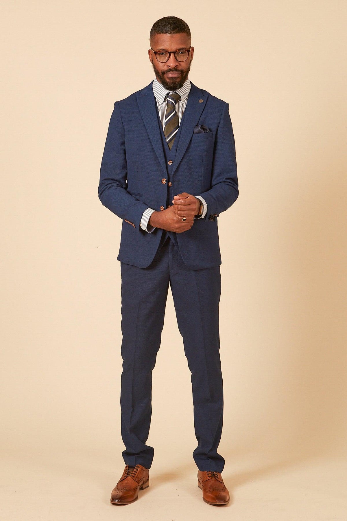 A man wears the marcdarcy Leicester City F.C. Collection - MAX Royal Blue Suit as worn by Harry Winks, paired with a striped shirt, patterned tie, brown dress shoes, and glasses, standing against a light beige background.
