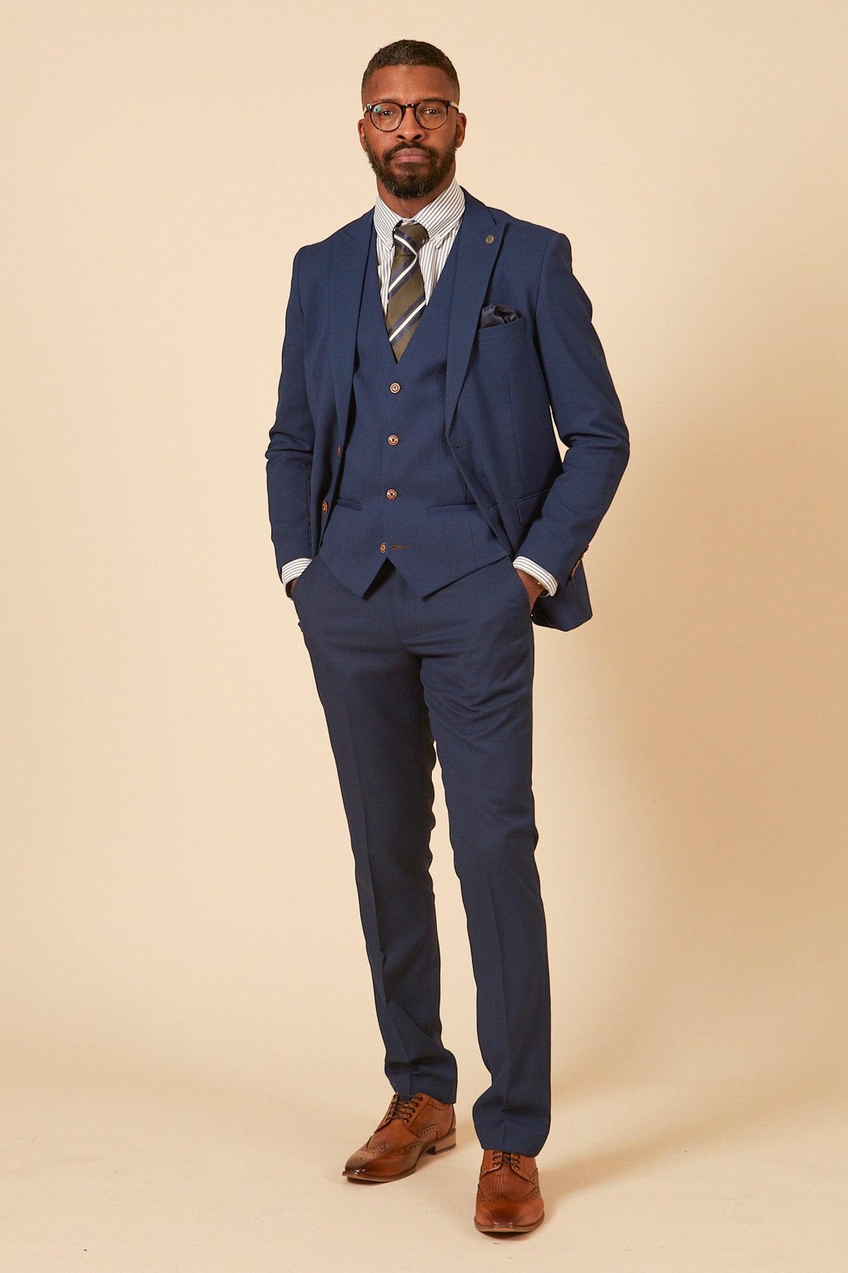 A Black man in a marcdarcy Official Leicester City F.C. Club Suit—MAX Royal Blue Suit with Kelvin Tan Waistcoat—wears a striped shirt and patterned tie, posing with his hands in his pockets.