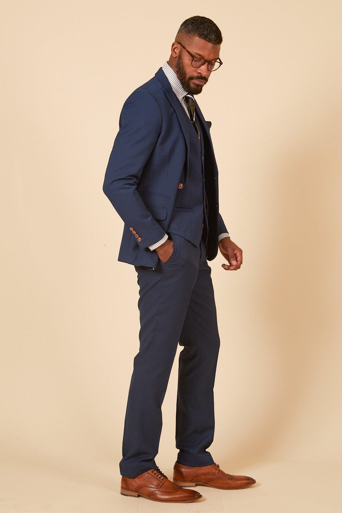A man stands in profile with one hand in his pocket, wearing the marcdarcy Official Cardiff City F.C. Club Suit | MAX Royal Blue Suit, paired with a vest, white shirt, tie, and brown dress shoes against a beige background.