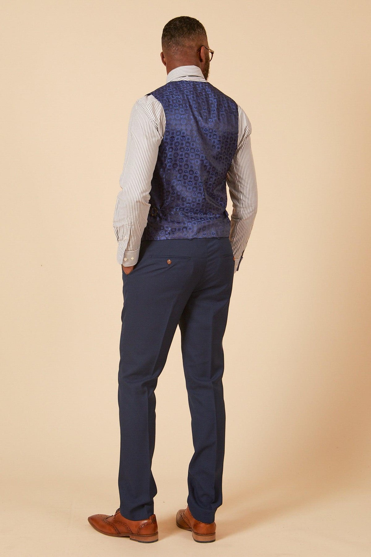 A man, facing away, wears the marcdarcy Leicester City F.C. Collection - MAX Royal Blue Suit as worn by Harry Winks, featuring a blue vest, striped shirt, slim-fit blazer, matching trousers, and brown dress shoes against a beige background.