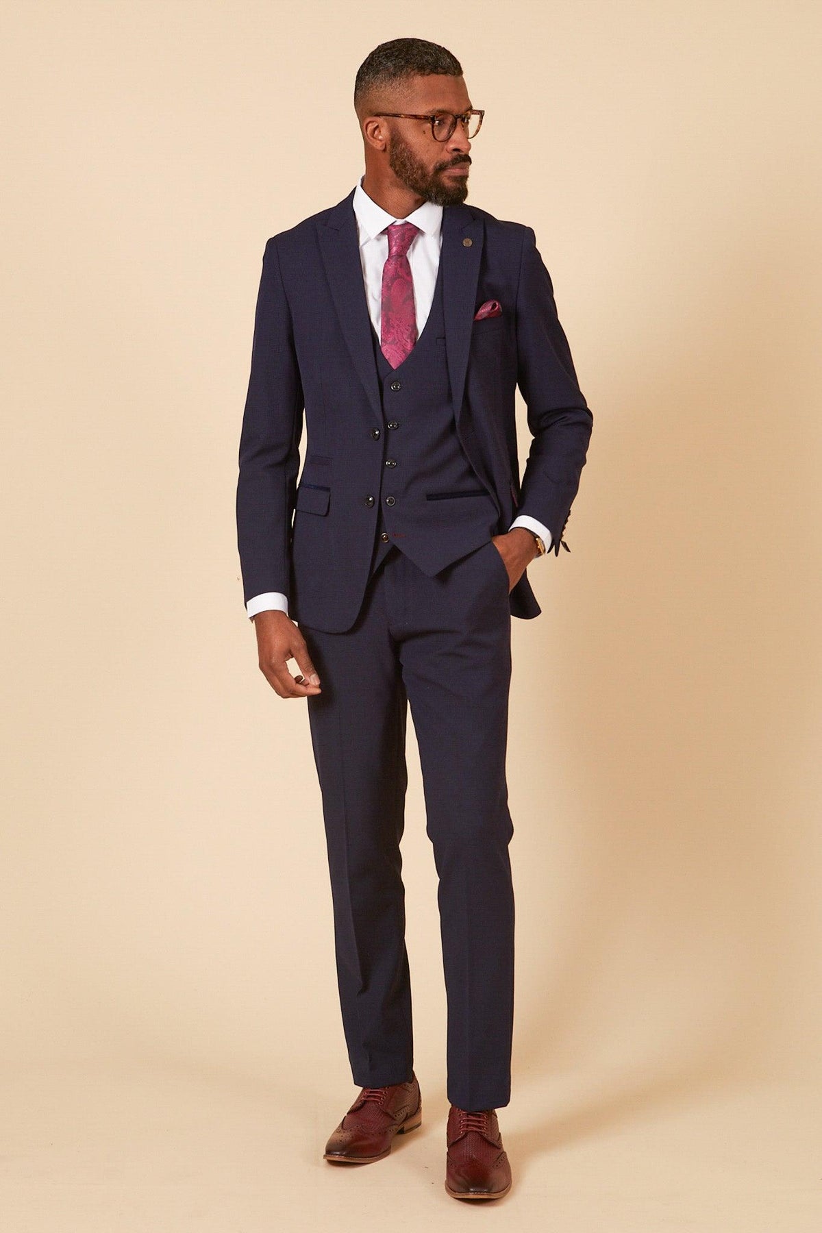 Lukasz Fabianski wears the Marc Darcy WHU Collection BROMLEY Navy Check Three Piece Suit, styled with a white shirt, pink paisley tie, matching pocket square, brown brogues, and tortoiseshell glasses against a beige backdrop.
