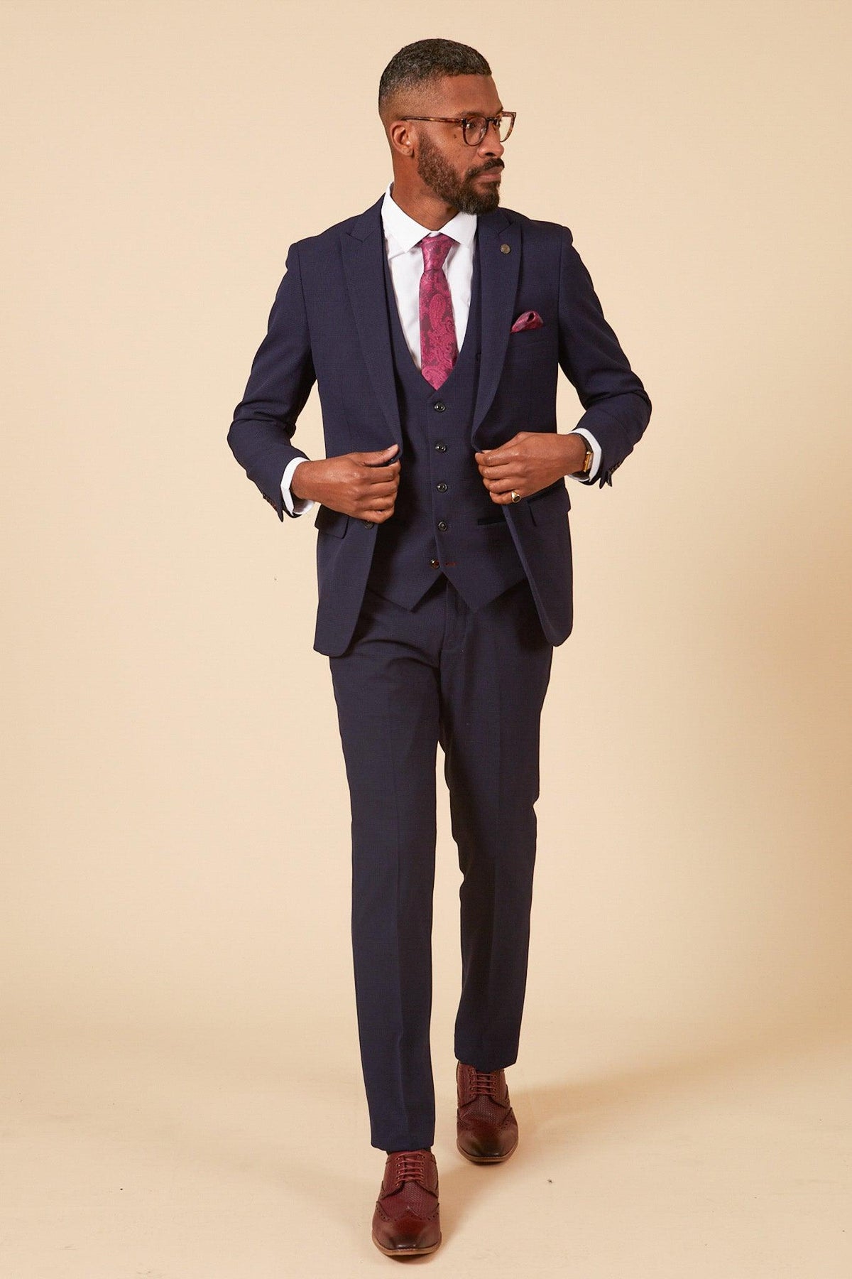 A man in Marc Darcys WHU Collection BROMLEY Navy Check Three-Piece Suit, as worn by Lukasz Fabianski, pairs it with a white shirt, burgundy tie and pocket square, and brown shoes while walking confidently against a beige background.