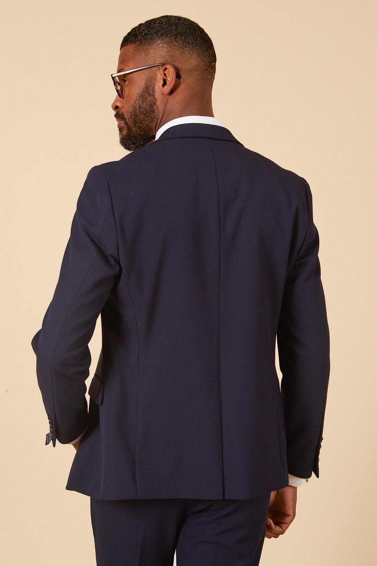 A man in the Marc Darcy WHU Collection BROMLEY Navy Check Three Piece Suit, as worn by Lukasz Fabianski, stands with his back to the camera and looks to the side against a plain beige background.