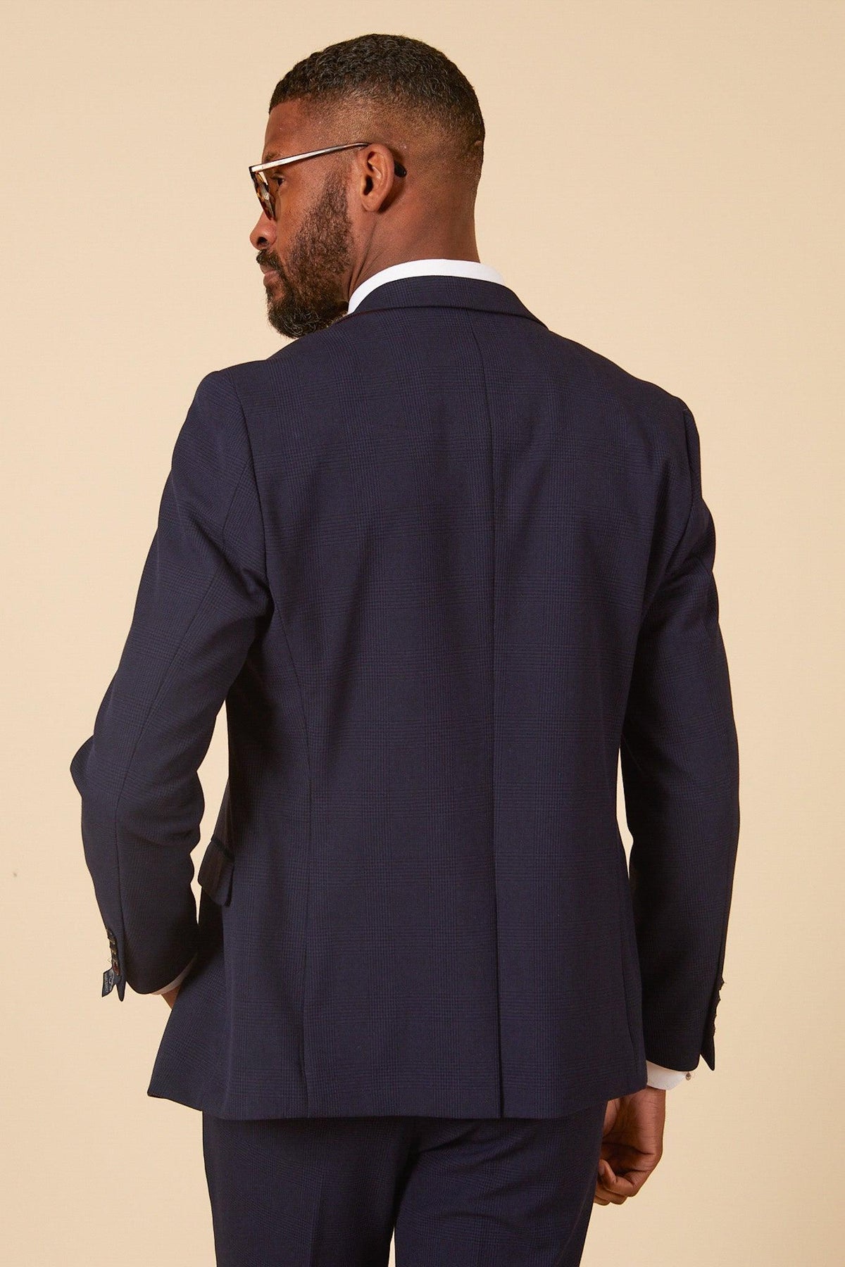 Back view of a man in the WHU Collection - BROMLEY Navy Check Three Piece Suit by Marc Darcy, as worn by Lukasz Fabianski, styled with a white shirt and matching trousers. He has a beard and sunglasses, set against a neutral beige background.