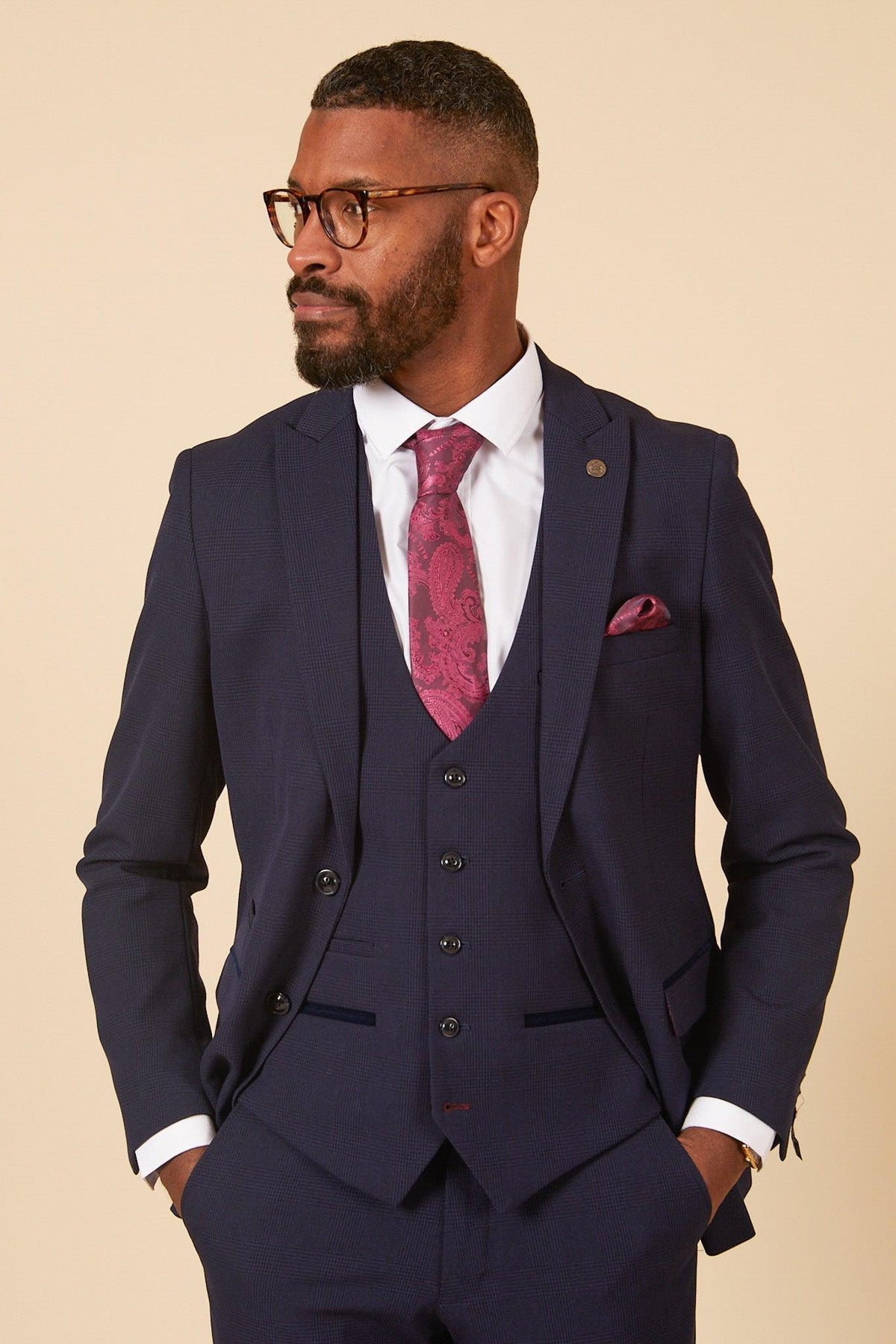 Man wearing The WHU Collection - BROMLEY Navy Check Three Piece Suit by Marc Darcy, paired with a white shirt, pink paisley tie, and pocket square. He has a beard, tortoiseshell glasses, hands in pockets against a beige background.