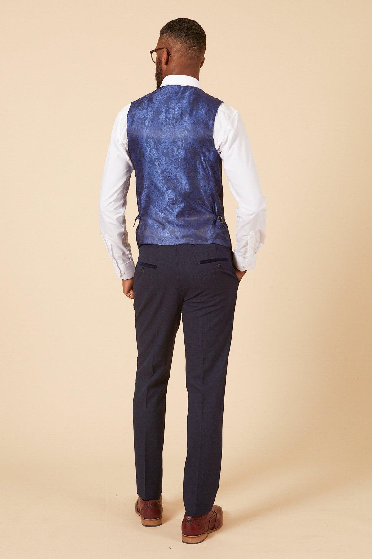 A man models the Marc Darcy WHU Collection - BROMLEY Navy Check Three Piece Suit as worn by Lukasz Fabianski, featuring a Prince of Wales check vest, white shirt, and navy pants, with brown shoes and glasses against a beige background.
