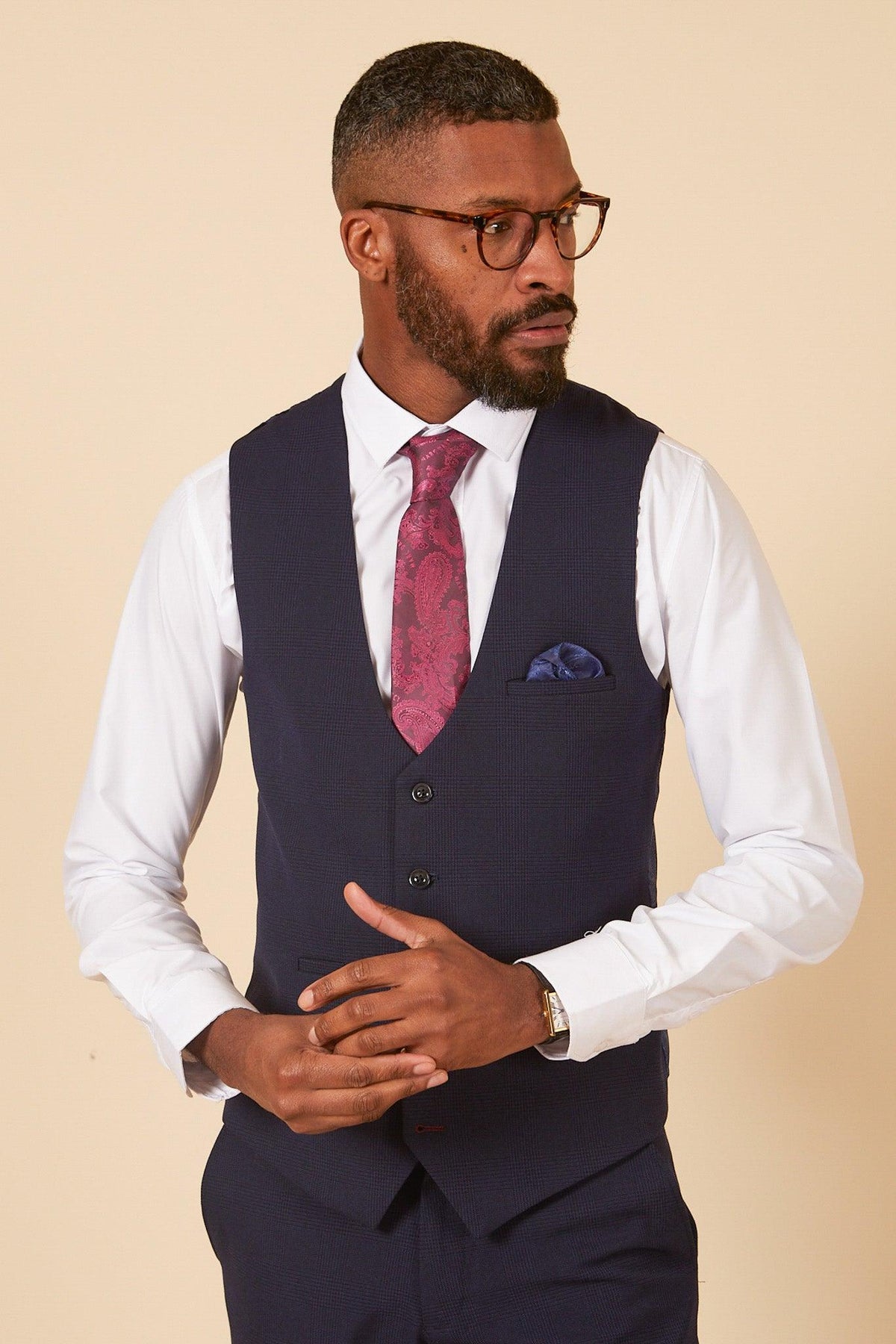 Lukasz Fabianski wears Marc Darcy’s WHU Collection - BROMLEY Navy Check Three Piece Suit, featuring a Prince of Wales check vest and maroon tie, with glasses and a white shirt, standing against a beige background.