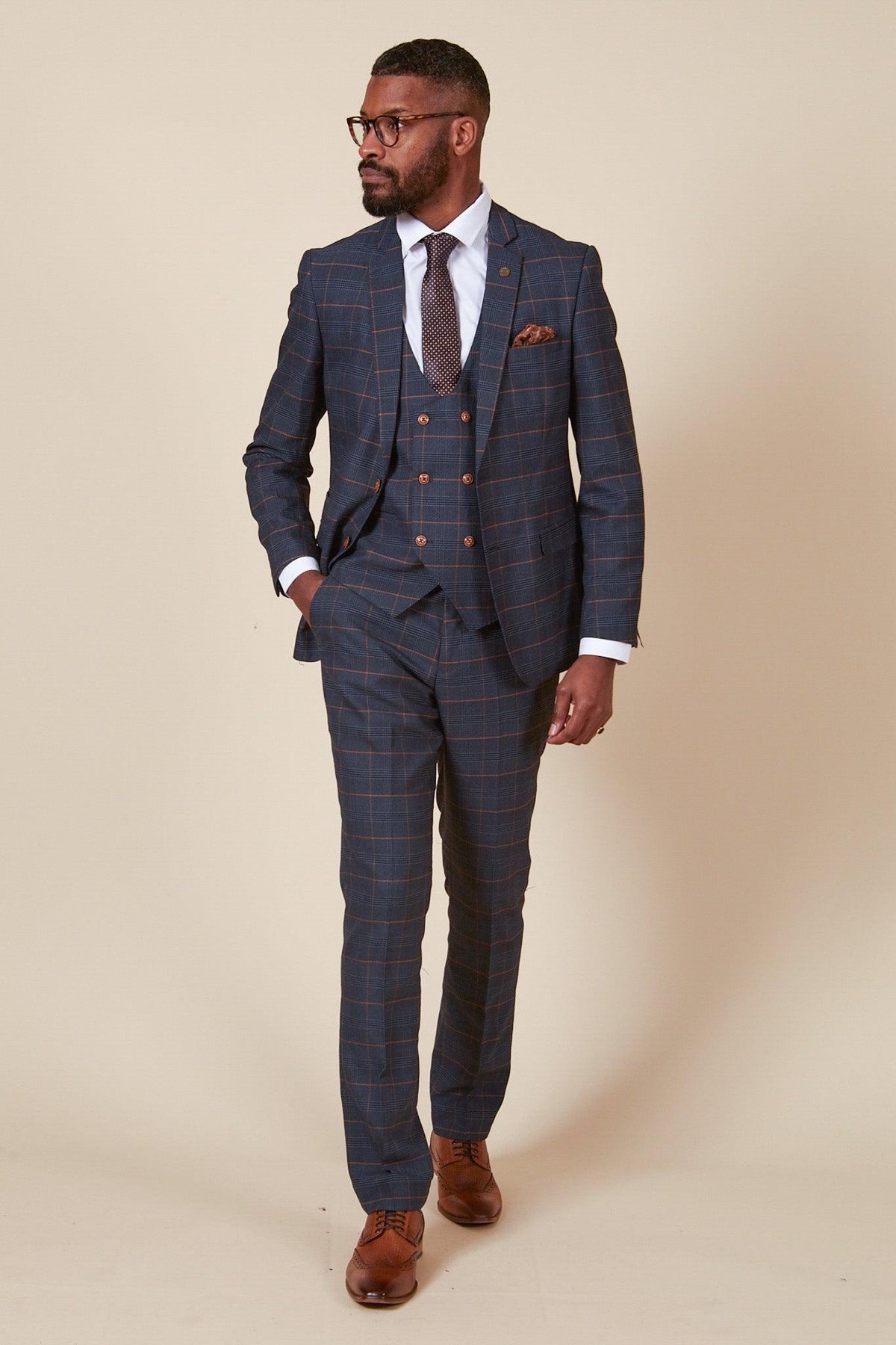 A man in the marcdarcy WBA Collection - JENSON Marine Navy Check Suit, as worn by John Swift, pairs it with a white shirt and patterned tie, standing confidently with his hand in his pocket.