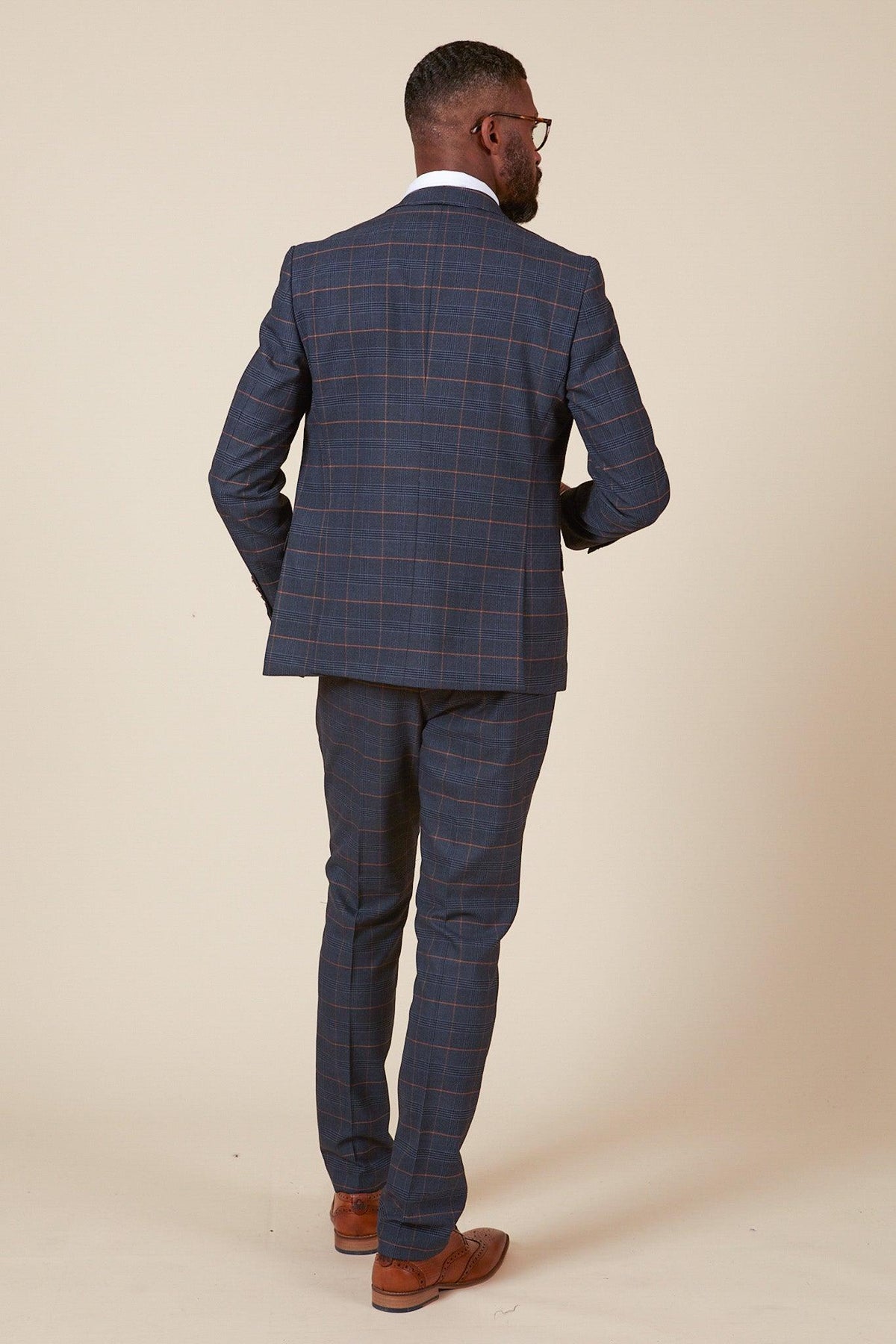 Man shown from behind wearing marcdarcy’s The WBA Collection - JENSON Marine Navy Check Suit As Worn By John Swift, featuring a slim-fit jacket and trousers, white shirt, tan brogue shoes, and glasses visible in side profile.
