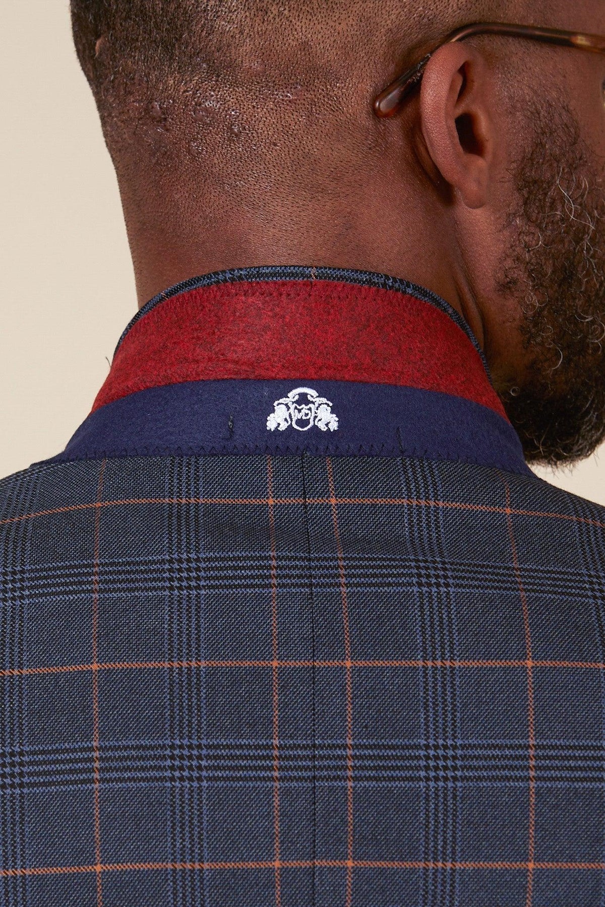 A close-up of a man's neck and shoulders wearing marcdarcy’s Jenson Marine Suit with Kelvin Tan Waistcoat from The WBA Collection, as worn by John Swift—featuring a red/navy collar and central white logo. Perfect for the Prince of Wales check style.