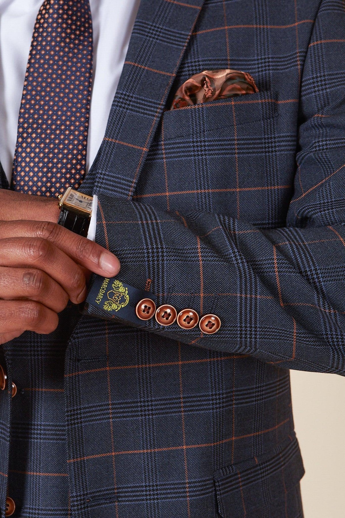 Close-up of the marcdarcy JENSON Marine Navy Check Suit from The WBA Collection, as worn by John Swift, featuring brown windowpane, copper buttons, bronze pocket square, paired with navy geometric tie and white shirt. Gold watch on wrist.