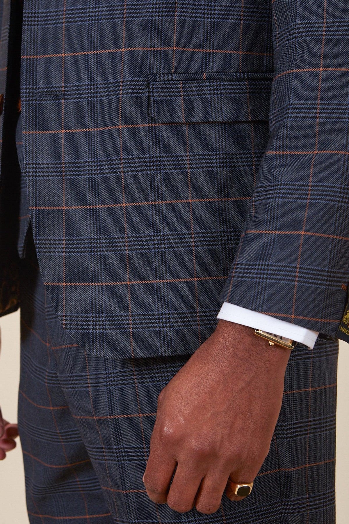 A person wears the marcdarcy WBA Collection - Jenson Marine Suit with Kelvin Tan Waistcoat as worn by John Swift, paired with a white shirt cuff, gold bracelet, and gold pinky ring. The image highlights their arm and torso.