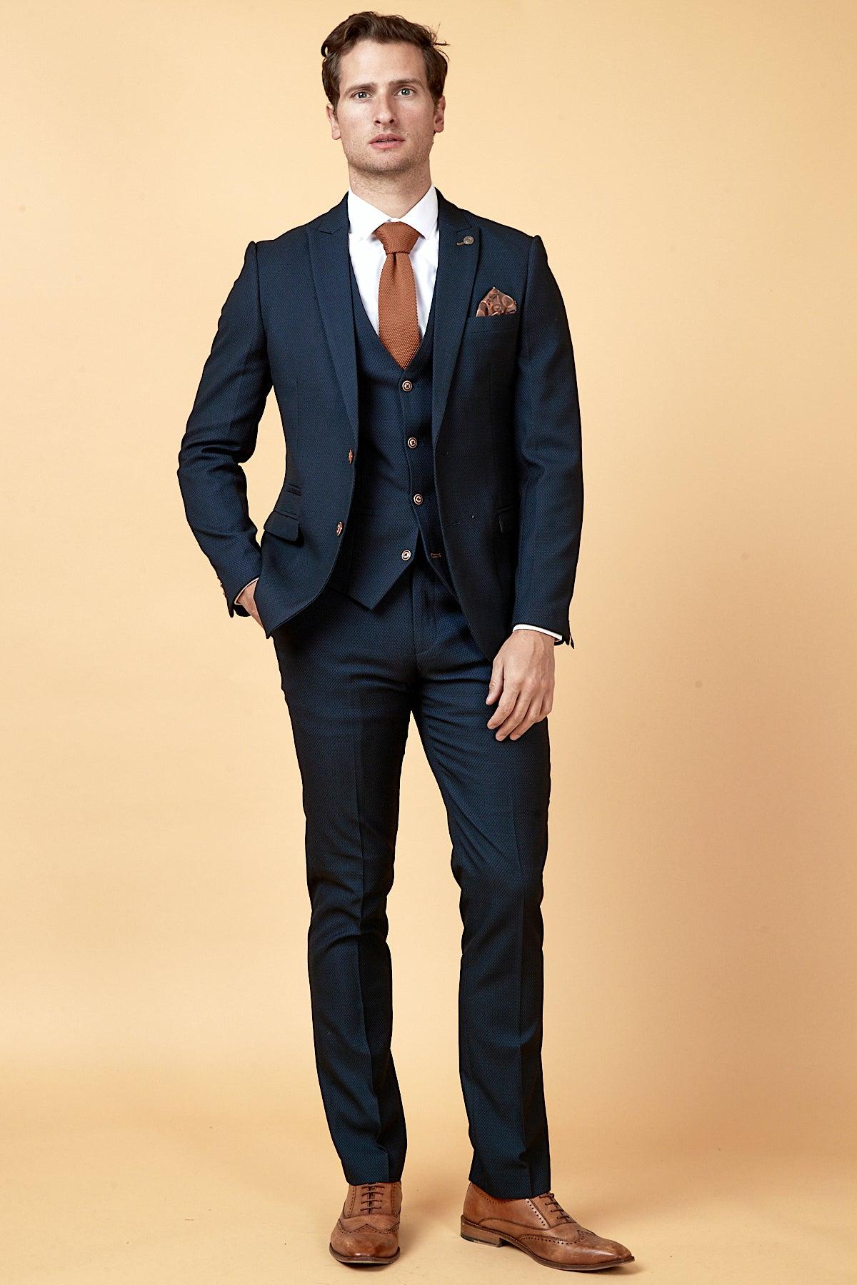 A man stands against a light beige background wearing the marcdarcy WHU Collection - MAX Navy Three Piece Suit as worn by Max Kilman, styled with a white shirt, tan tie, brown pocket square, and brown dress shoes.
