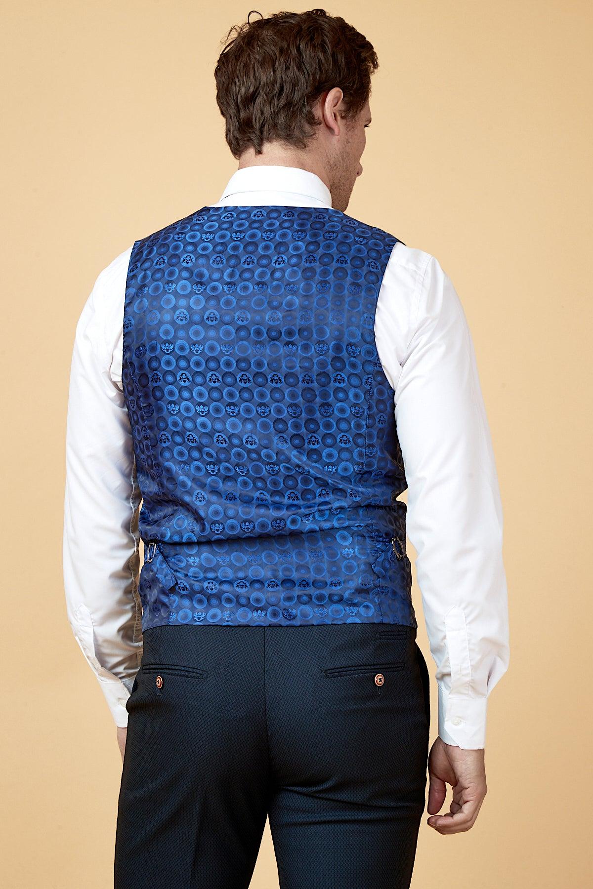 A man faces away wearing the marcdarcy WHU Collection - MAX Navy Three Piece Suit, as worn by Max Kilman, featuring a white shirt and blue vest with circular patterns, styled against a plain beige background.