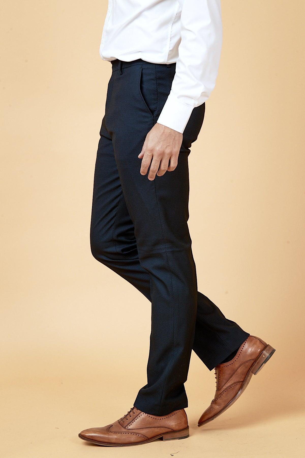 A man wears the marcdarcy WHU Collection MAX Navy Three Piece Suit, as worn by Max Kilman, with a white dress shirt and brown leather wingtip shoes, standing against a tan background.