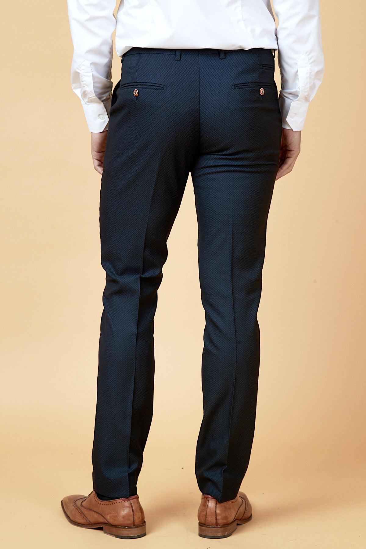 A person is seen from behind against a beige background, wearing marcdarcy's The WHU Collection - MAX Navy Three Piece Suit As Worn By Max Kilman, paired with a white dress shirt and brown dress shoes.