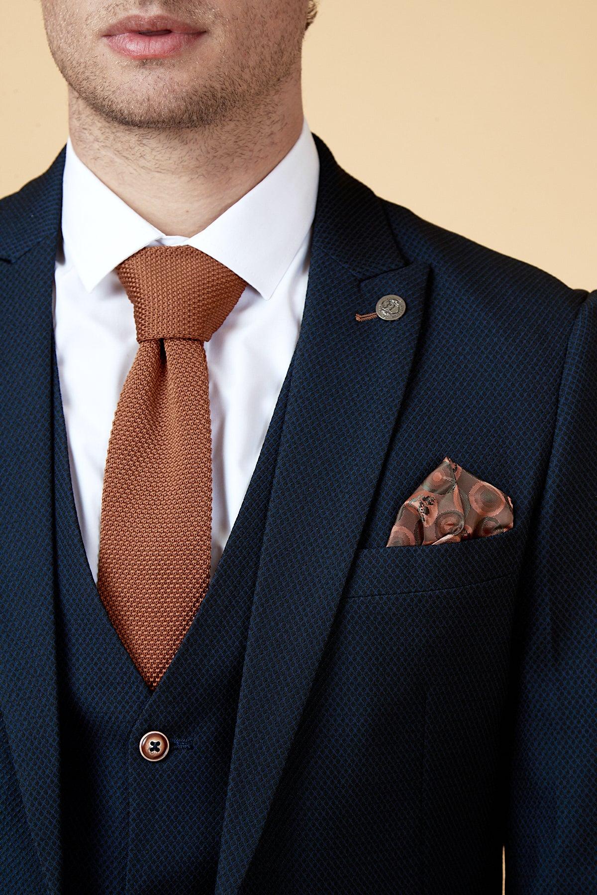 Close-up of a man wearing the marcdarcy WHU Collection - MAX Navy Three Piece Suit as worn by Max Kilman, styled with a white shirt, brown knitted tie, patterned pocket square, and lapel pin.