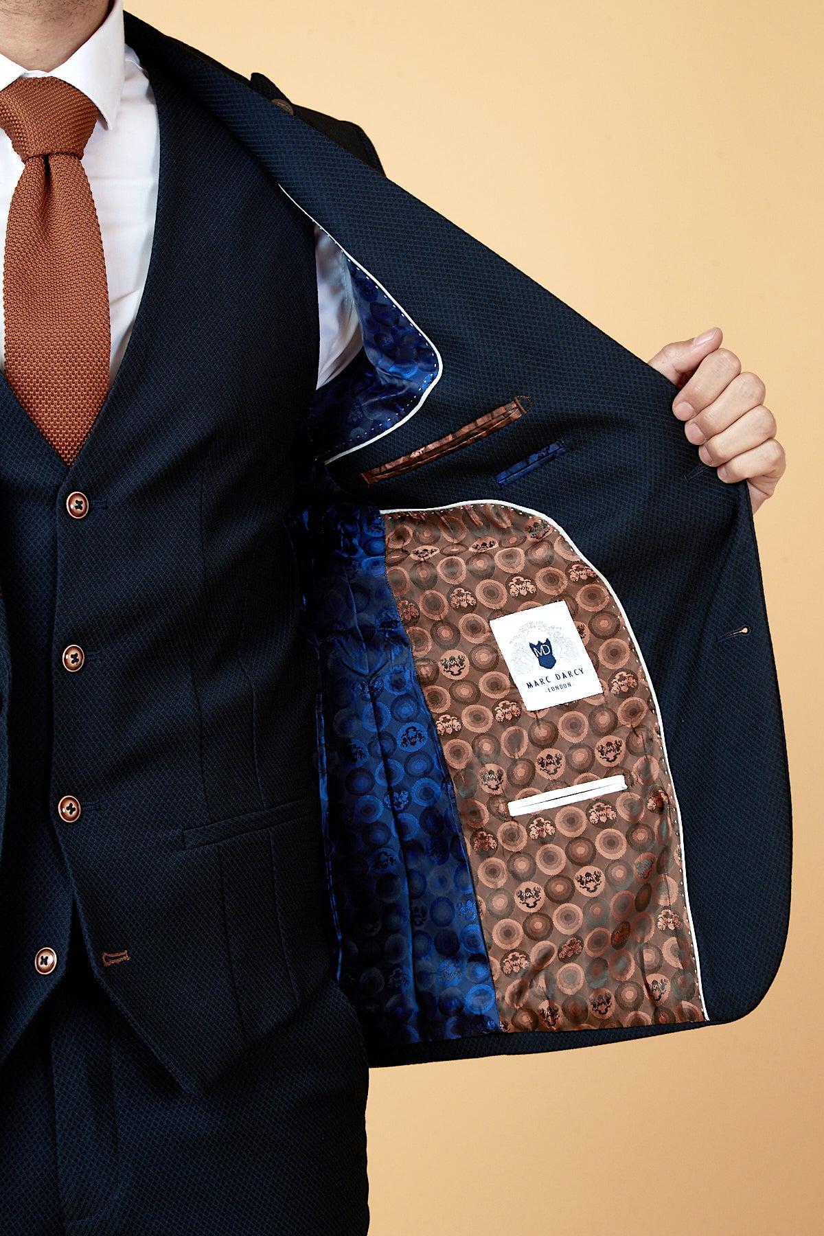 A man wears the marcdarcy WHU Collection - MAX Navy Three Piece Suit as worn by Max Kilman, paired with a white shirt and orange tie. The open slim fit jacket reveals its patterned blue and brown lining with the Marc Darcy London label.