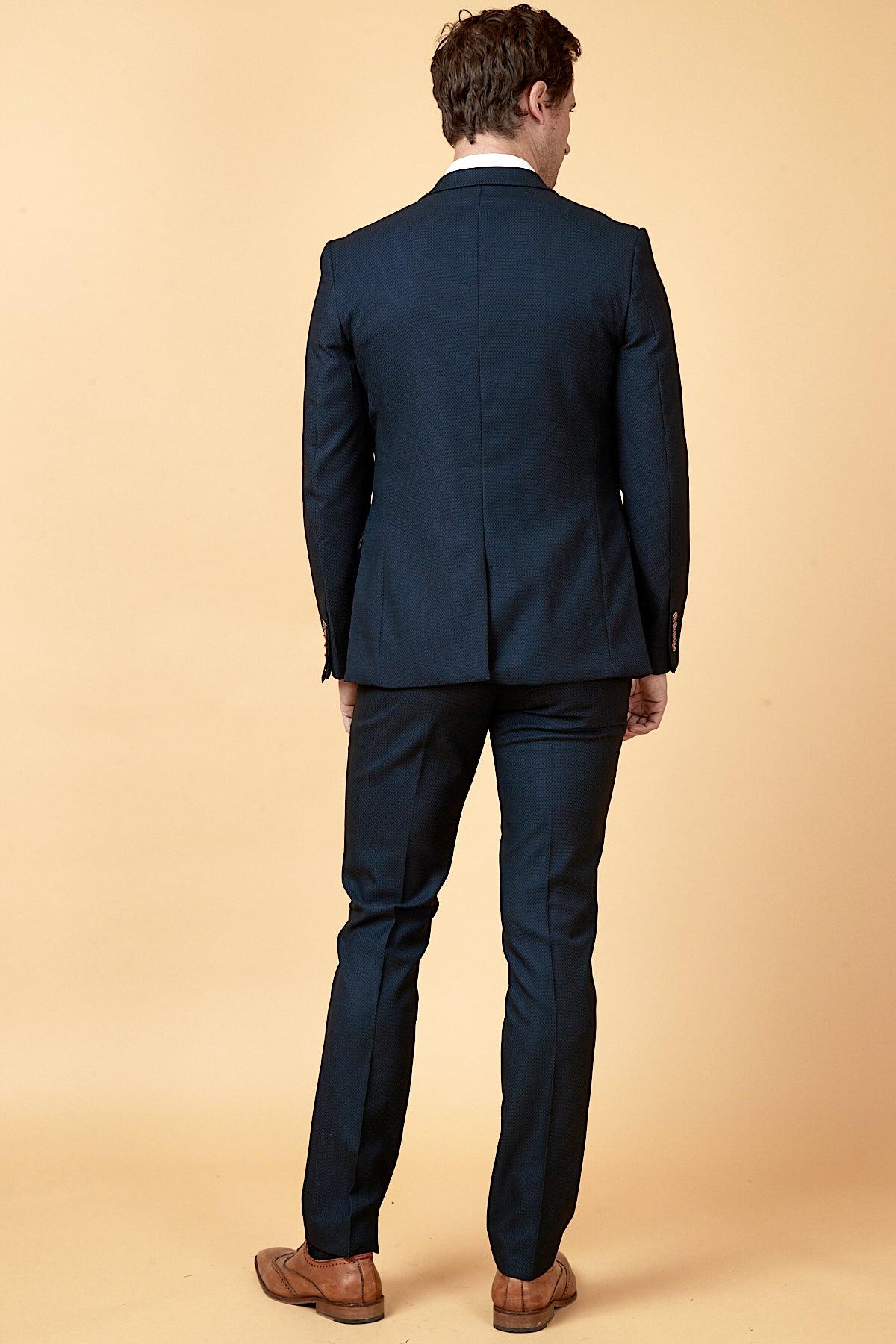 Back view of a man wearing marcdarcy's WHU Collection - MAX Navy Three Piece Suit as worn by Max Kilman, featuring subtle patterns and brown dress shoes, standing against a cream background.